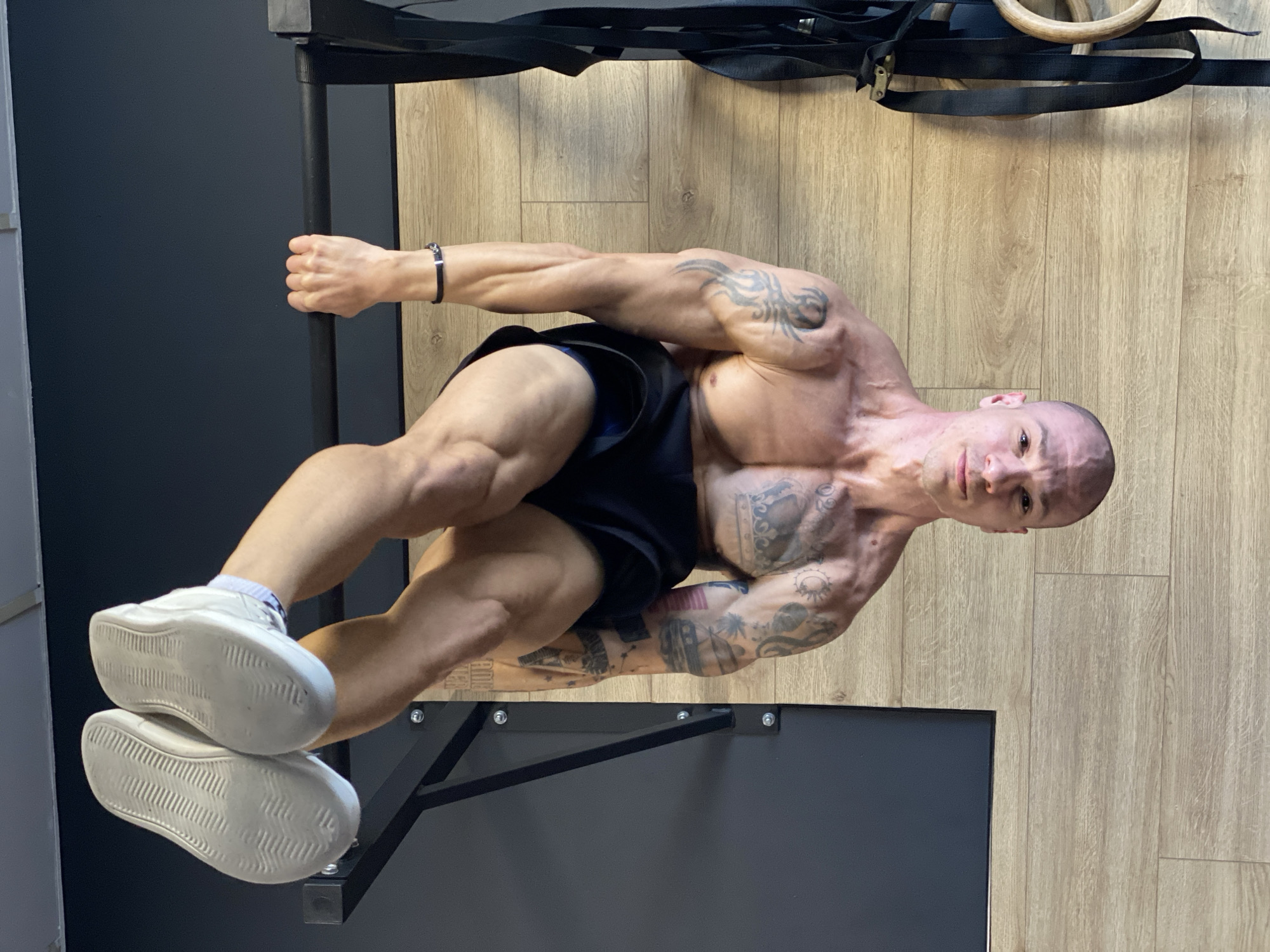 A muscular man with tattoos is performing a calisthenics exercise in what appears to be a gym setting. He is holding onto a black metal bar with one hand and resting his feet on a black horizontal structure. He is wearing black shorts and white sneakers with visible white socks. Above him, there are black straps and wooden gymnastic rings. The background consists of a light-colored wood floor and a dark-gray wall. The location is not discernible as being in Pleven, Bulgaria, based on the visible content of the image.