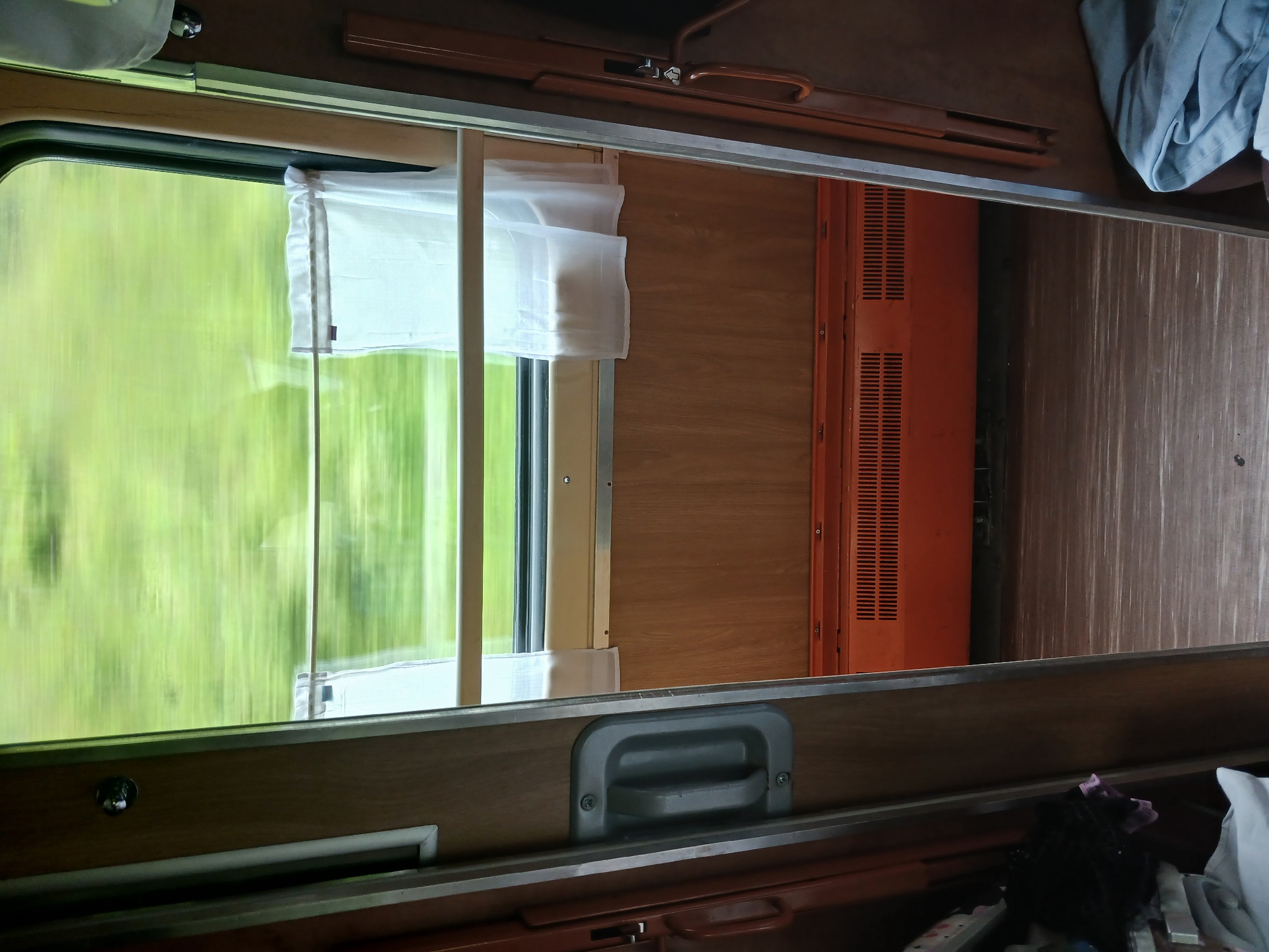 This is a photograph taken from inside a train carriage, looking out through an open doorway towards the window. The scene is one of movement, as indicated by the blurred green foliage seen through the window, suggesting the train is traveling through a natural landscape, likely rural or mountainous. The interior of the carriage is simple and functional, with wooden paneling on the walls and a wooden floor. A gray handle is visible on the left doorframe, and a brown handle on the right. An orange heating unit with a slatted front is mounted near the floor beneath the window. Two sheer white curtains hang on a rod across the lower part of the window. On the left, near the open doorway, a pile of items including dark fabric and a pink cloth are visible. On the right, a folded light blue fabric is present. The overall impression is that of a basic, utilitarian train compartment. The image does not contain any elements that would specifically place it in Krasnaya Polyana, Russia.