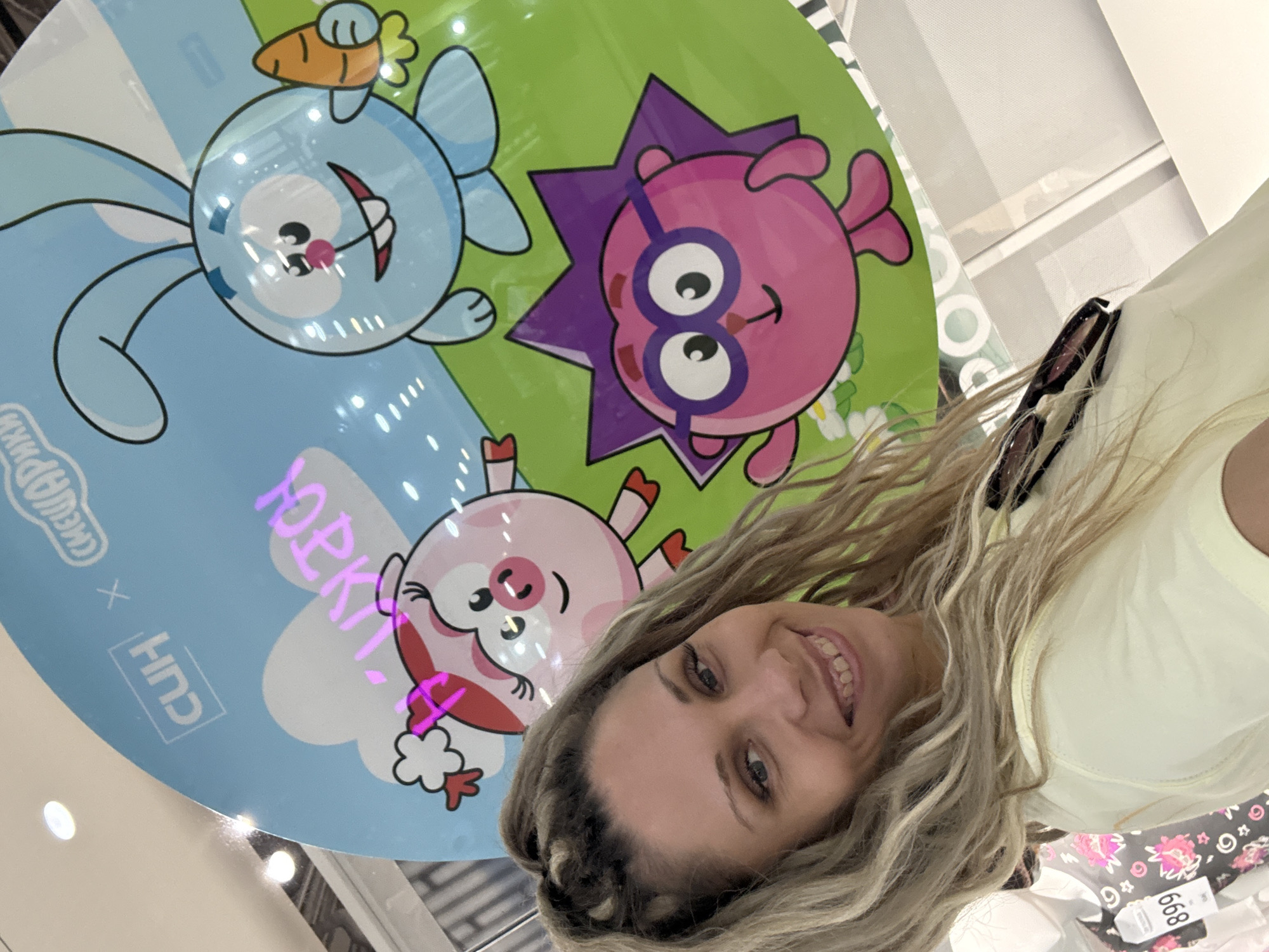 A blonde woman is taking a selfie in front of a large, circular, light blue display featuring cartoon characters, possibly a promotional event. The characters are a blue rabbit with a carrot, a pink pig with a bow, and a pink hedgehog wearing glasses. The text on the display includes "CUH x СМЕШАРИКИ" in white and pink lettering. The woman is smiling and wearing a light yellow shirt. In the background, there are mannequins with clothing, one with a tag labeled "899". The environment appears to be indoors, possibly a shopping mall or store. There is no indication this scene is located in Sosnovtsy, Russia.