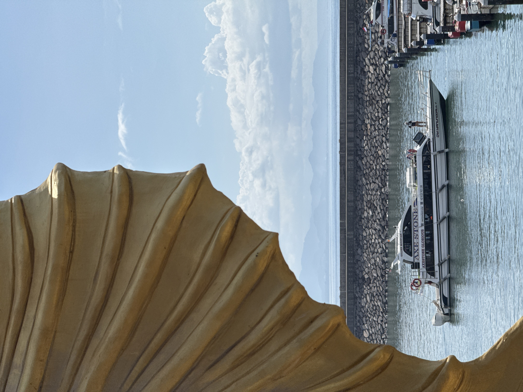 The image depicts a scenic view in Denpasar, Indonesia. In the foreground, there is a large, textured, golden-colored structure, possibly part of a sculpture or architectural element. Beyond that, there is a body of water, likely a harbor or bay, with several boats docked. One of the boats, named "AXE STONE," is prominently featured, with the text "AXESTONE GLORIOUS" and a phone number (+62 8123 7621 130) visible on its side. There appears to be at least one person on board. The background includes a rocky shoreline, a clear sky with scattered clouds, and distant mountains.