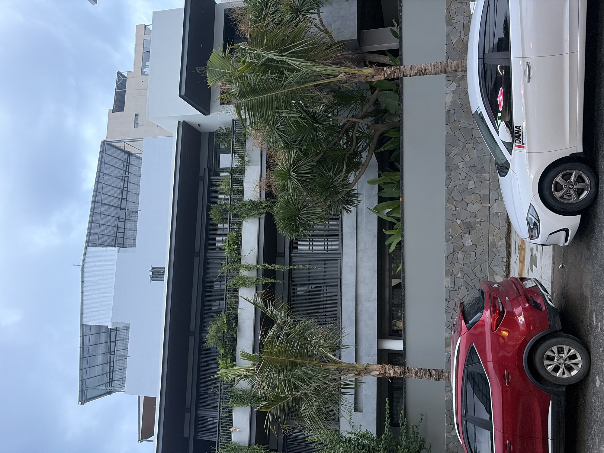 Two cars, a red MG ZS and a white Hyundai Elantra, are parked on a street in front of a modern building with multiple levels and balconies. The building features a gray facade, dark-framed windows, and a tiered roof structure with metal awnings. Lush greenery, including palm trees and hanging plants, surrounds the building. The street appears wet, suggesting recent rain. The white car has "DANA" branding visible on its side. The scene is captured on a cloudy day.