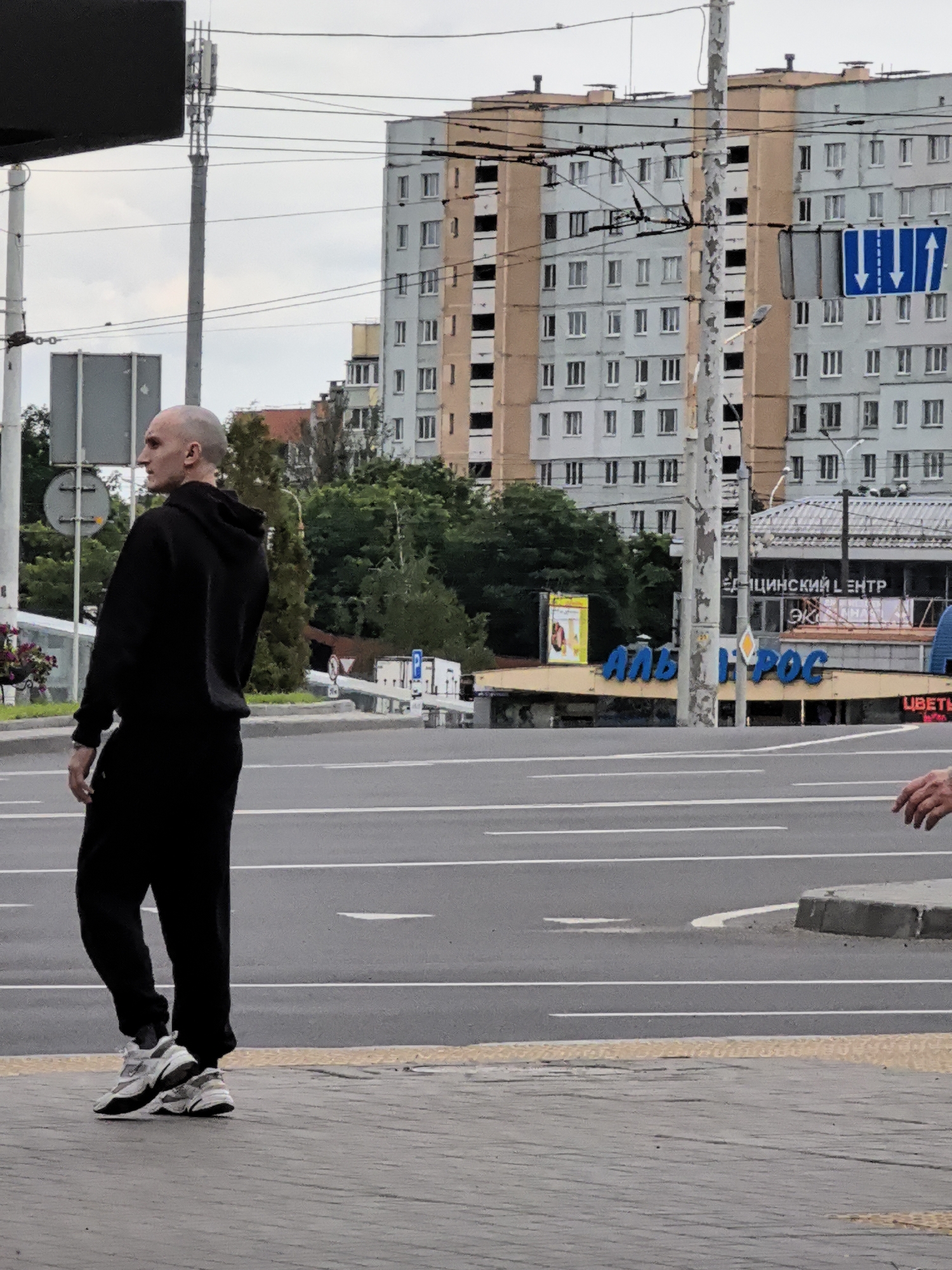 A bald man in a black hoodie and black pants stands on a sidewalk in front of a street. He is facing to the left, looking towards the street. In the background, there are buildings, trees, and traffic lights. There are also some power lines running across the image. The sky is overcast.