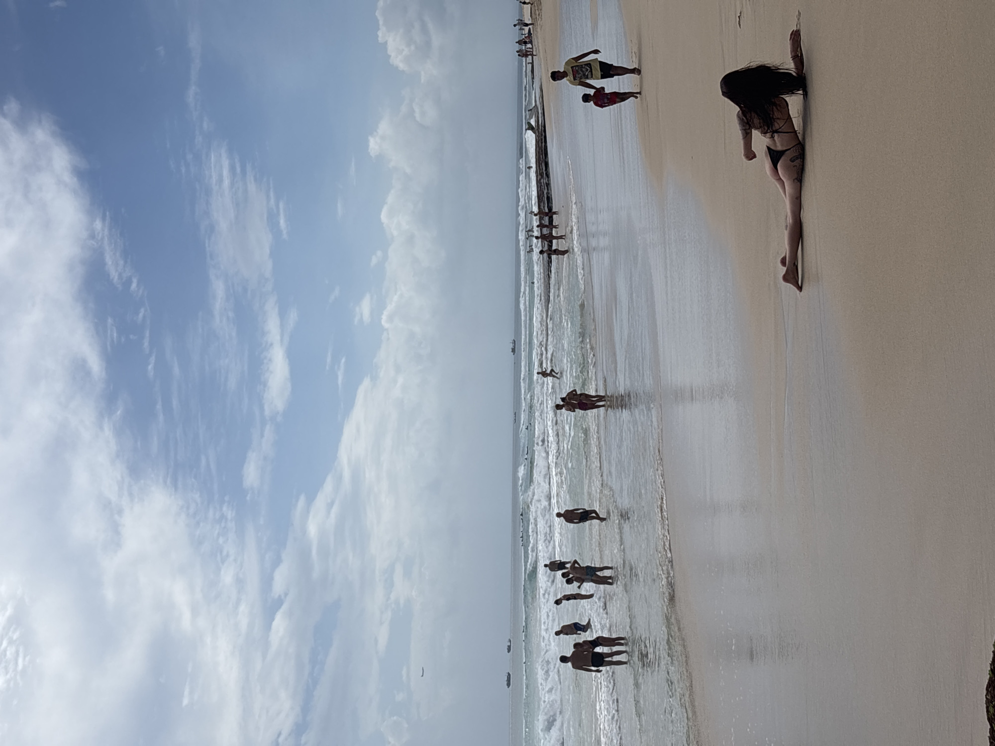The image depicts a sunny day at a beach in Kuta Selatan, Indonesia. The sky is filled with scattered white clouds against a blue backdrop. The ocean is calm with gentle waves breaking on the shore. Numerous people are visible in the water and on the sand, engaging in various activities. Some are wading in the shallow water, while others are swimming or enjoying the waves. A few individuals are seen walking along the shoreline. In the foreground, a person is lying on the wet sand near the water's edge, facing away from the camera and towards the ocean. In the distance, several boats are anchored, and an airplane can be seen flying in the sky. The beach is sandy with some rocky outcrops visible in the water.
