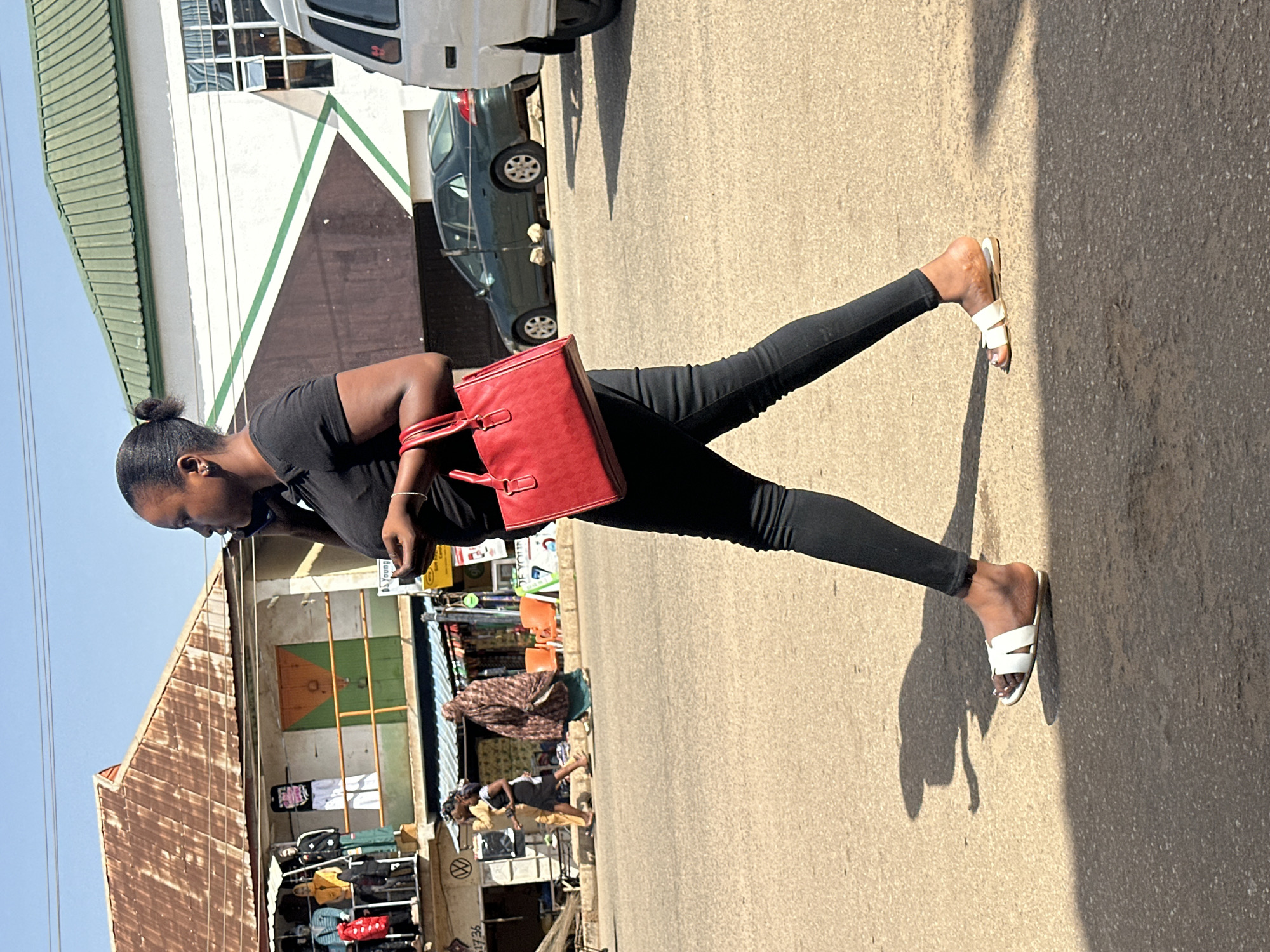 A young Black woman walks on a paved street in Bukuru, Nigeria, holding a red handbag and talking on her phone. She wears a black t-shirt and black leggings, with white slides on her feet. Sunlight casts a shadow of her on the street. In the background, shops with corrugated metal roofs line the street. A green sedan and a white van are parked on the side of the road. Other pedestrians can be seen walking in the distance.