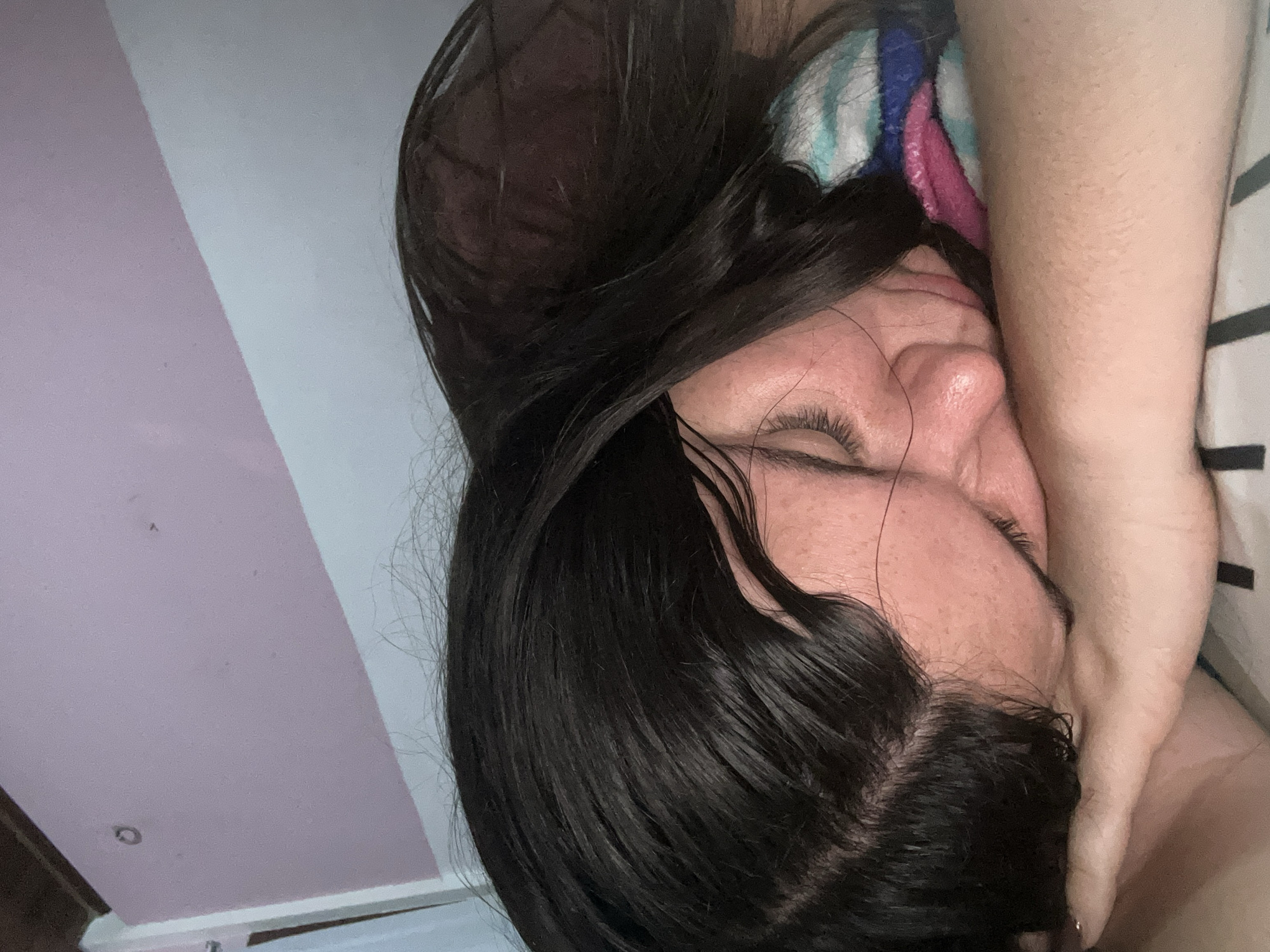 A close-up, tilted, indoor shot depicts a woman with dark hair, her eyes closed. Her face is pressed against her hand/arm, which is also covering part of her face. A multicolored striped object (possibly a pillow or towel) is behind her head. The background consists of a wall with vertical stripes of white and light pink/purple. No information is available to confirm the location is San Juan de Los Morros, Venezuela.