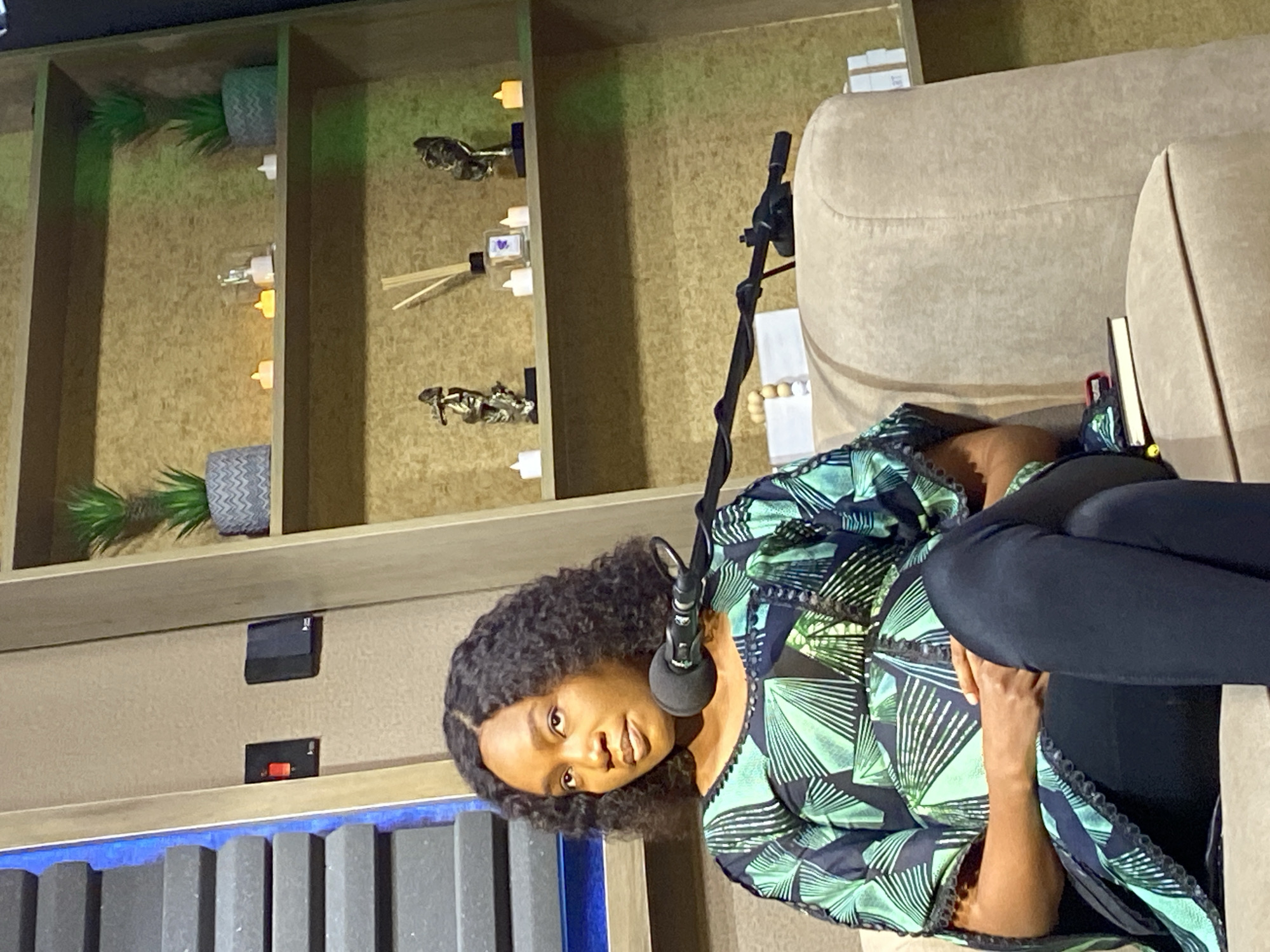 The image shows a woman sitting on a beige couch in what appears to be a recording studio, given the presence of a microphone in front of her. She has dark, curly hair and is wearing a patterned green and black dress. Her posture suggests she is engaged in a conversation or presentation.
Behind her, a wall with shelves is visible, decorated with artificial plants in patterned pots, small decorative figurines, and what look like scented diffuser sticks. There are also small, faux candles or LED lights on the shelves, adding a warm ambiance. In the lower part of the frame, a black object with a red switch is mounted on the wall, possibly a power outlet or a control panel. A section of soundproofing material is also visible at the bottom of the image.
The lighting appears to be artificial, with a slight green tint in some areas, possibly from studio lighting. The overall scene suggests a professional setting for interviews, podcasts, or broadcasts. No visible text is present in the image.