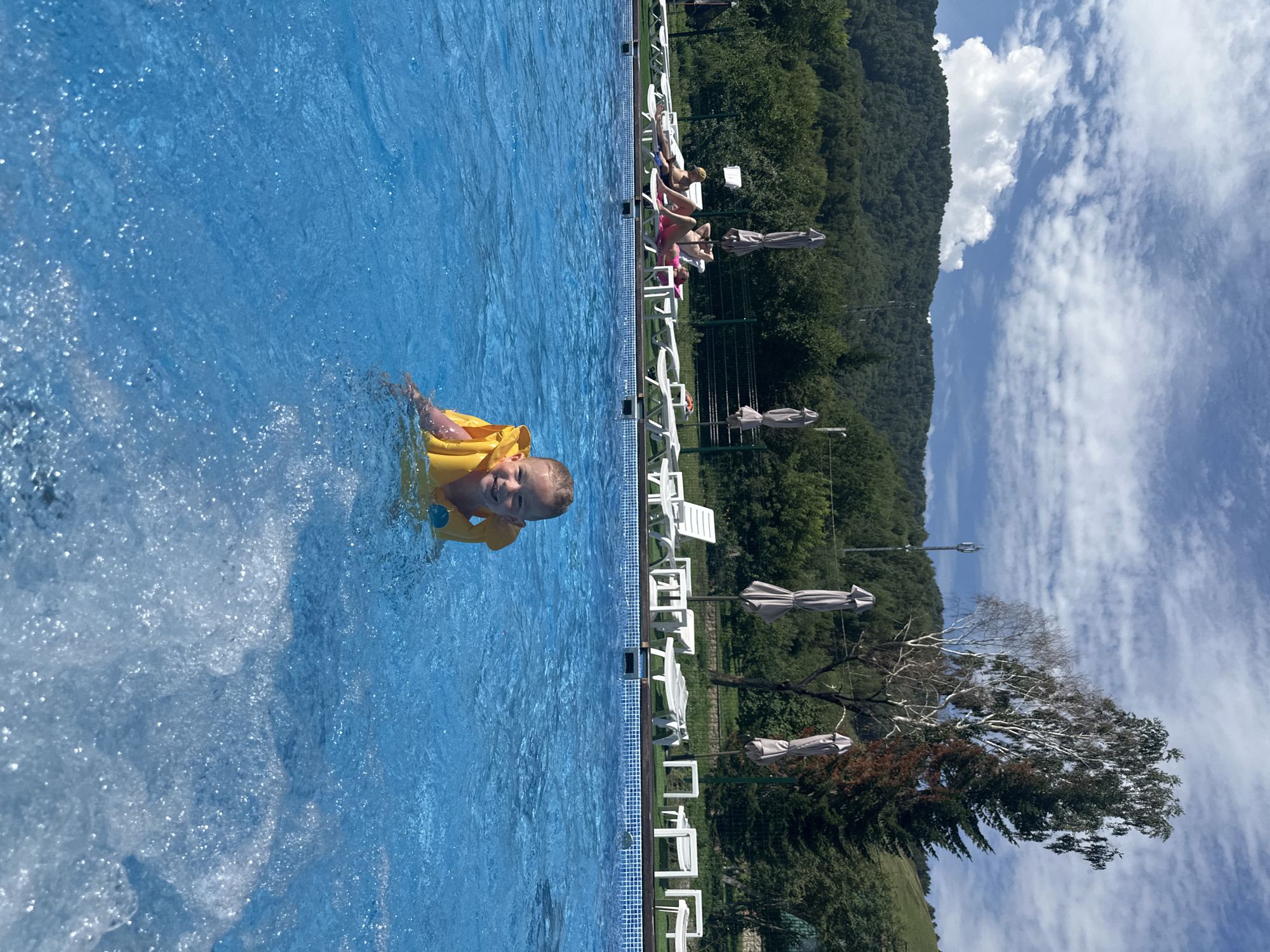 A young girl wearing a yellow life vest is swimming in a bright blue outdoor swimming pool on a sunny day. The pool is surrounded by lounge chairs and umbrellas, with a lush green forest and rolling hills in the background. The sky is partly cloudy with dramatic white clouds against a blue expanse. In the distance, a cell tower is visible on a hilltop. The scene appears to be a resort or recreational area, likely in the vicinity of Даниловка (алтайский), Russia, given the natural landscape.