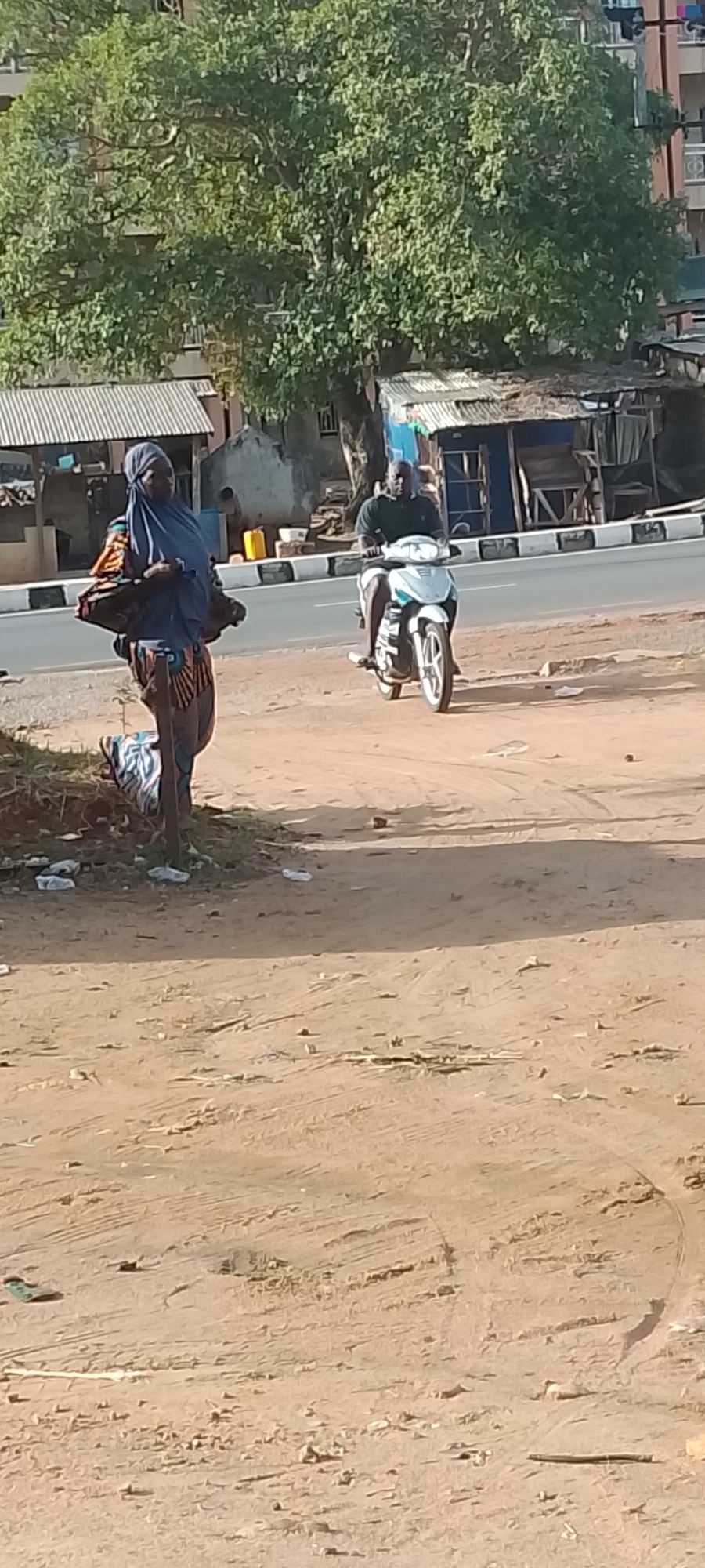 The image shows a street scene in Adankolo, Nigeria. A woman in a blue hijab and colorful patterned clothing stands on a dirt patch, possibly a roadside or undeveloped area. She is carrying something across her body, obscuring her view. Adjacent to the dirt patch is a road with black and white striped edges. A man is riding a white and turquoise scooter on the road. In the background, there are small, makeshift structures or stalls along the road. Beyond that, there is a large tree and a taller building. The environment appears to be sunny.