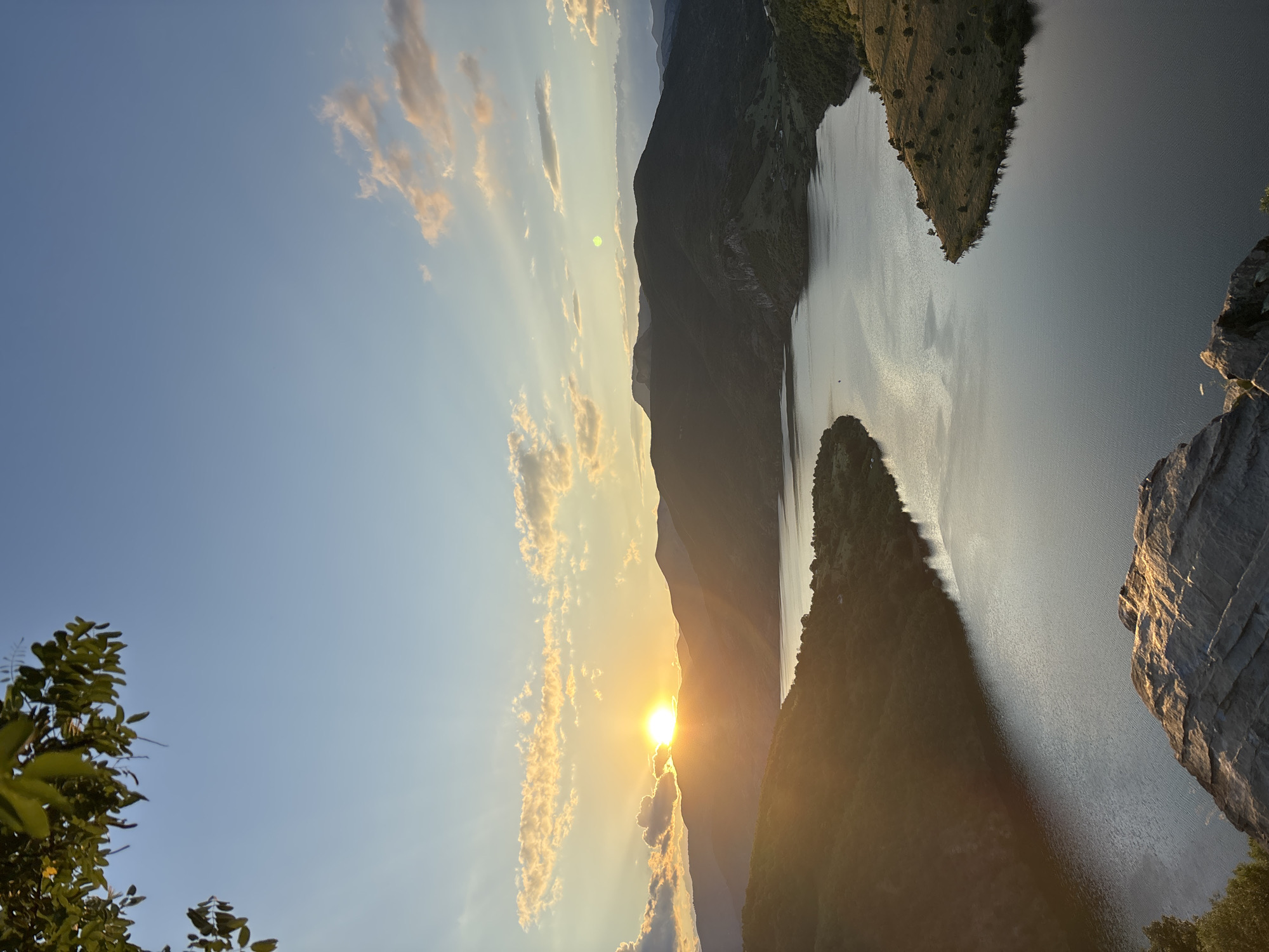 This image, taken in the vicinity of Borovitsa, Bulgaria, captures a serene natural landscape at sunset. A vast, calm lake dominates the central and lower portions of the frame, its surface reflecting the warm hues of the setting sun. Surrounding the lake are rolling hills and mountains covered in lush greenery. In the foreground, a rocky outcrop with textured surfaces is visible, illuminated by the golden light. The sky above is a gradient of soft blues and oranges, with scattered clouds tinged by the sunset. The sun, a bright orb, is positioned behind the distant mountains on the left, casting long shadows and creating sunrays that pierce through the atmosphere. There are no people or animals visible in the image. The overall scene conveys a sense of tranquility and natural beauty.