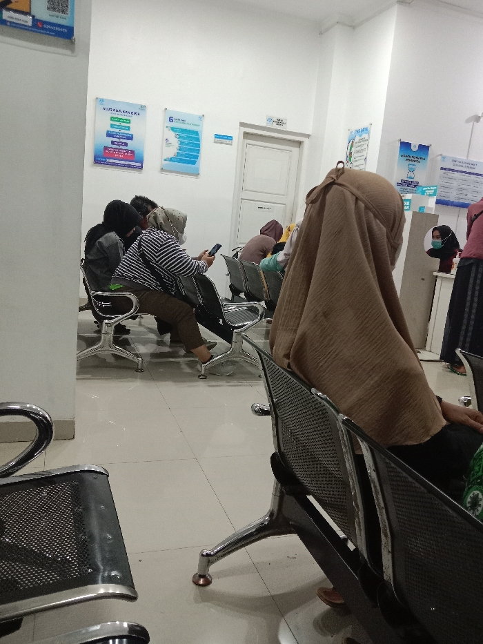 An indoor scene in a waiting area in Gladaksari, Indonesia, depicts approximately six to eight individuals seated in rows of connected waiting chairs with chrome frames and black mesh seats. The walls are white, and the floor is a reflective, light-colored tile. Several informational posters with blue and white designs are affixed to the walls; one poster on the left displays the number "6" and another has a QR code. A light-colored door with horizontal panels is visible in the background, beneath a small sign.
In the immediate foreground, facing away from the viewer, is the back of a person wearing a light brown hijab and dark clothing, seated on a black and chrome chair. To their left, in the midground, a person wearing a striped long-sleeve shirt and dark trousers is seated, looking down at a black smartphone held in both hands. This individual is also wearing a light-colored head covering or mask. Adjacent to them, another person in a black hijab and dark top is seated. Further back, additional individuals are visible, also seated, including one wearing a dark reddish-brown hijab and another with a dark blue face mask partially visible behind a white counter-like structure on the far right. No direct interactions are occurring; individuals are passively waiting.