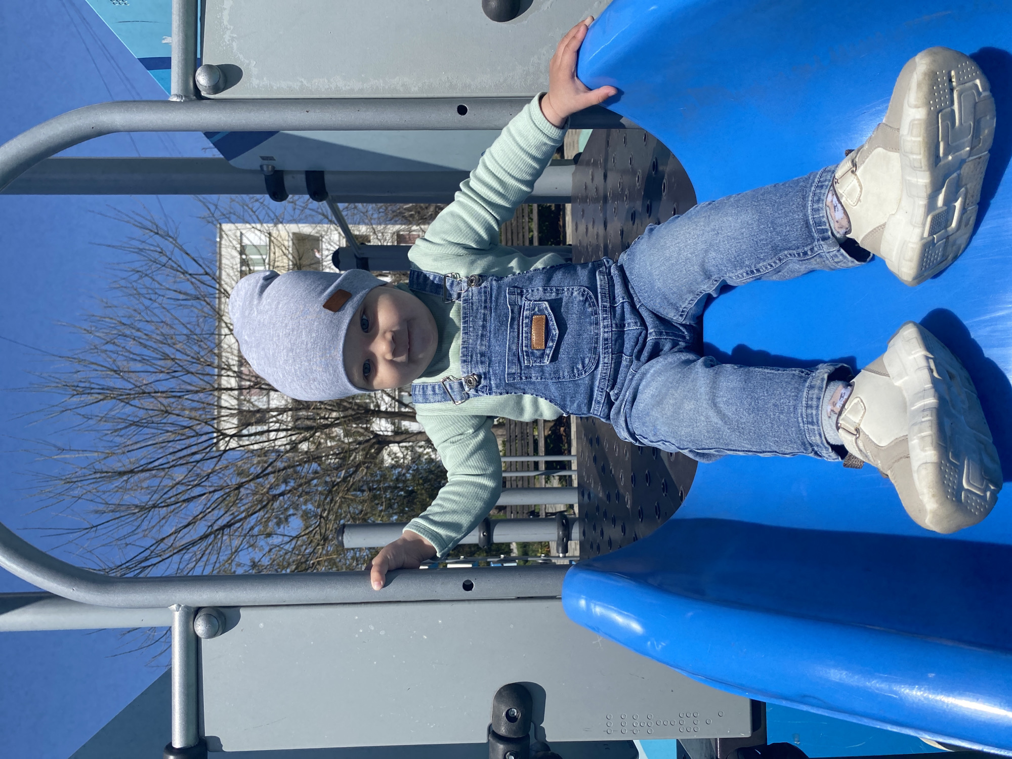 A young child in a grey hat and denim overalls sits on a blue playground slide. The child wears a light green long-sleeved shirt and beige shoes. The playground features grey metal bars and structures around the slide. Bare trees are visible in the background against a clear blue sky, along with a light-colored building. The location is possibly in Sevastopol, Ukraine, based on the prompt's mention.