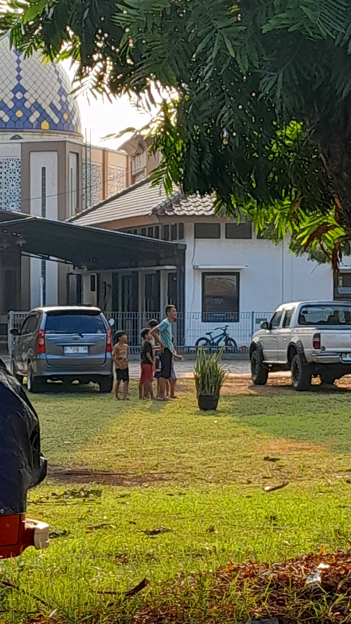 In the city of Unknown, Unknown, the image depicts an outdoor scene with several individuals and vehicles. In the foreground, a patch of green grass is visible, with some scattered leaves and debris. A dark object, possibly a piece of furniture or a vehicle component, is partially visible on the left.
The middle ground features a group of people, including an adult male and four children, standing on the grass. The adult appears to be wearing a light blue t-shirt and dark shorts. The children are dressed in casual clothing. Behind them, a silver minivan is parked. Further to the right, a potted plant with tall, green foliage stands next to a silver pickup truck. A bicycle is leaning against a fence in the background, near a white building with a tiled roof. A large tree with dark green leaves is positioned in the upper right corner of the frame, partially obscuring the sky and a dome-shaped structure with blue and white patterns. Sunlight filters through the leaves, creating a dappled effect. The overall environment suggests a residential or communal area.