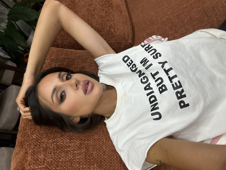 An image taken in Khimki, Russia, depicts a female individual lying on an orange-brown, textured fabric sofa. The individual, appearing to be in her 20s or 30s, has dark hair and wears full facial makeup, including winged eyeliner and light pink lipstick. She is dressed in a white, short-sleeved t-shirt with cut-off sleeves, bearing the black text "UNDIAGNOSED BUT I'M PRETTY SURE" across the chest. Faint discoloration is visible on the upper left chest area of the t-shirt. Her right arm is raised, with her hand positioned near her head; a ring with a clear stone is visible on her right ring finger. A small, horizontal mark or scar is present on her right upper arm. On her left forearm, a faint tattoo reading "measure" is discernible. Her expression is direct, facing the camera. The background reveals an indoor setting with a portion of light-colored furniture and a green potted plant visible behind the subject.