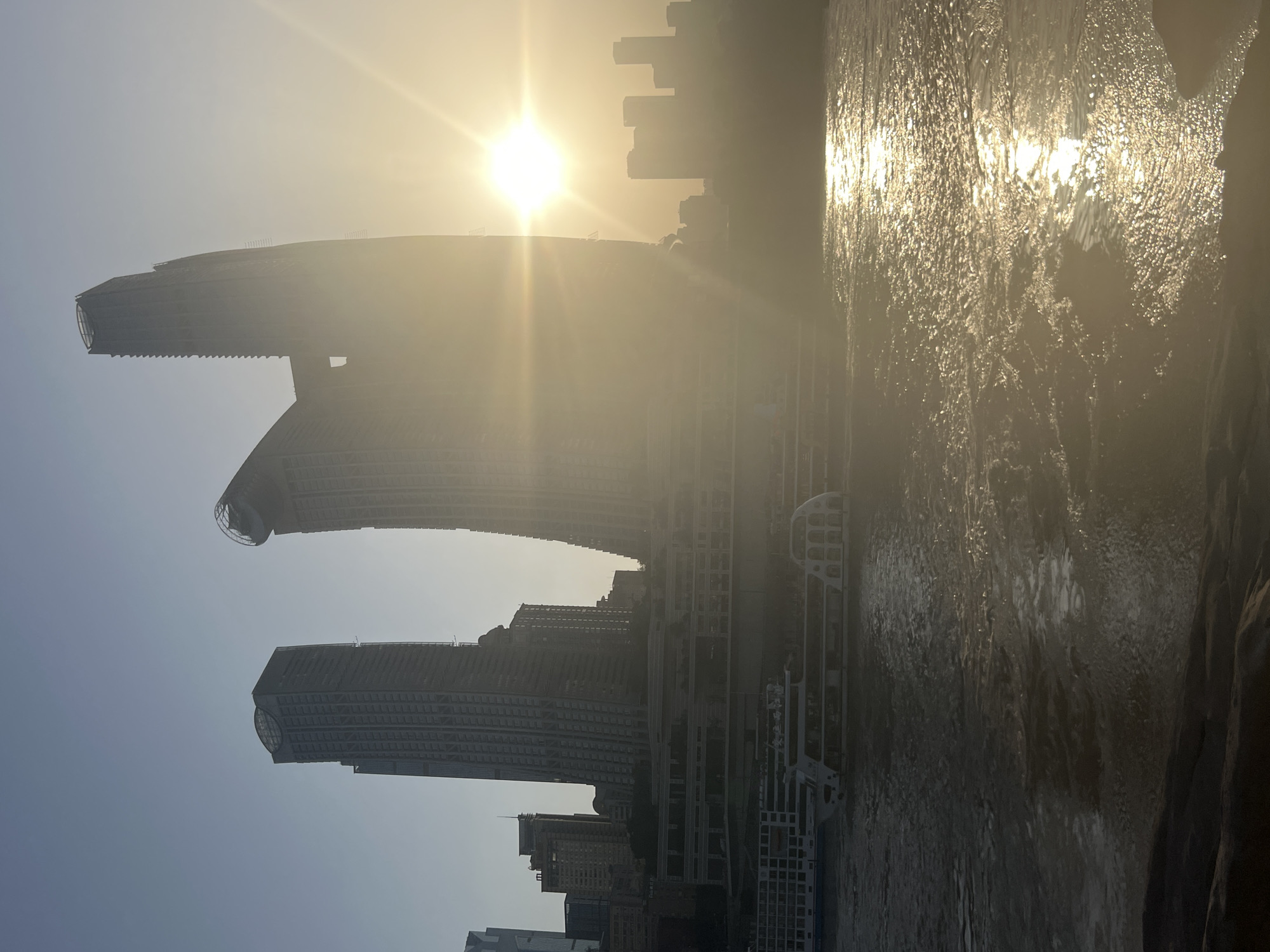This is a wide-angle, eye-level shot of the Yangtze River at sunset in Nan'an District, China. The sun is low in the sky, casting a warm, hazy glow over the scene. In the foreground, the river is choppy and reflects the sunlight, creating a shimmering effect. Several large tour boats are docked or moving slowly on the water. In the mid-ground, a multi-level waterfront complex with a modern, curved architectural design is visible. Behind this, a series of tall, contemporary skyscrapers dominate the skyline, with two particularly distinct buildings in the center right of the frame, one appearing to be under construction. The sky is a clear, pale blue, fading into a hazy yellow near the sun.