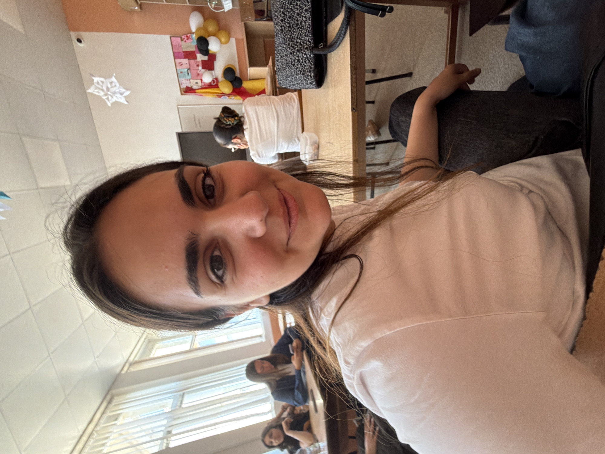 The image is a selfie of a woman with long dark hair, fair skin, and brown eyes, wearing a light pink or white top. She is looking directly at the camera with a slight smile.
In the background, several people are seated at desks in what appears to be a classroom or a similar setting, with windows visible on the left side of the frame, suggesting a well-lit interior. Some decorative items can be seen on the back wall, including balloons and a star-shaped ornament, indicating a festive or celebratory occasion.
Based on the metadata, the image was taken in Burgas, Bulgaria.