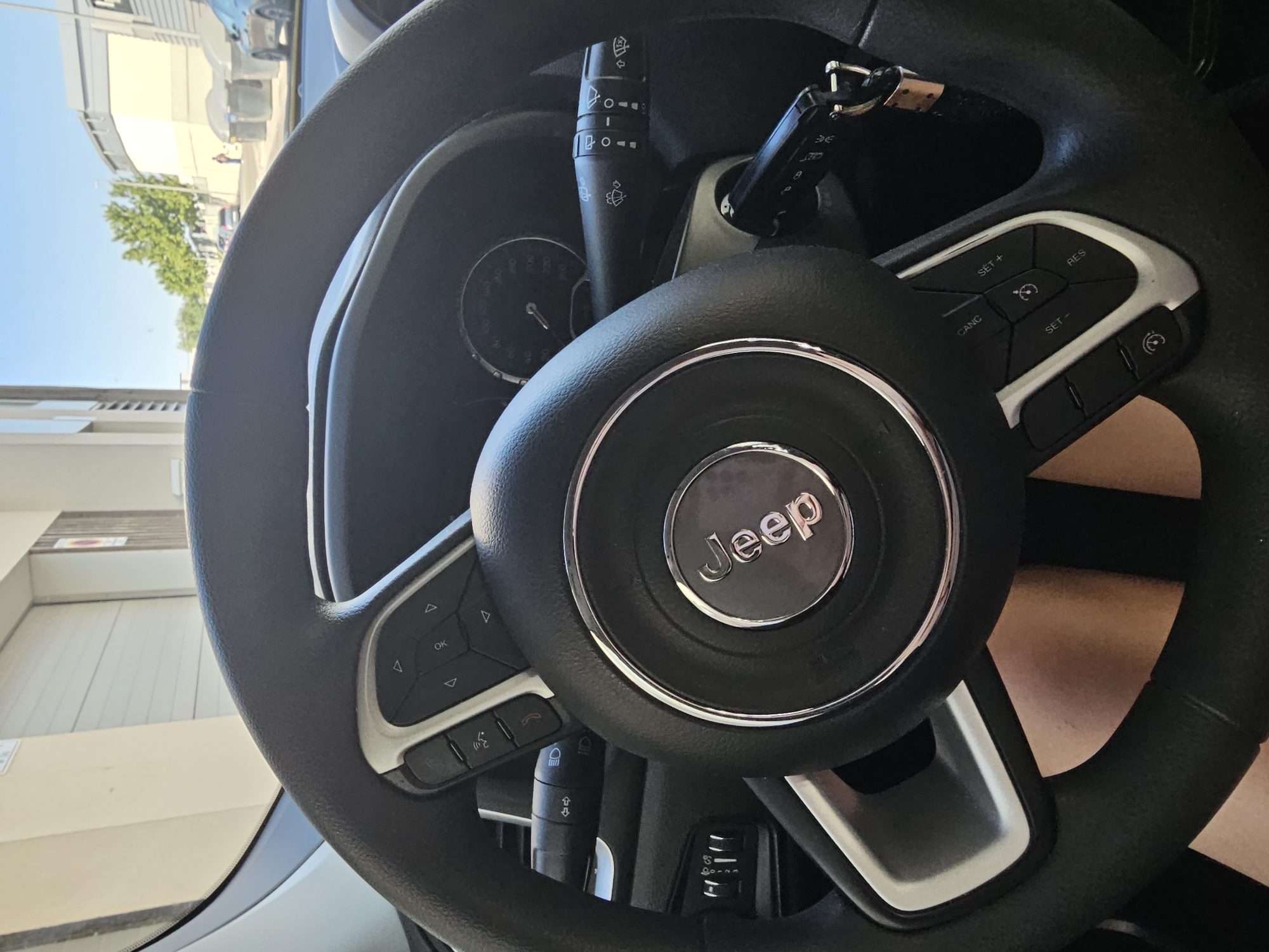 The image shows a close-up view of a black Jeep steering wheel, with the Jeep logo prominently displayed in the center. The steering wheel has various buttons on either side, including controls for the audio system, cruise control, and potentially other vehicle functions. A car key is inserted into the ignition on the right side of the steering column. The background is slightly out of focus, but suggests an outdoor setting with a building and trees visible. Given the context of the location, it is highly probable that this image was taken within the city of Cuarte de Huerva, Spain.