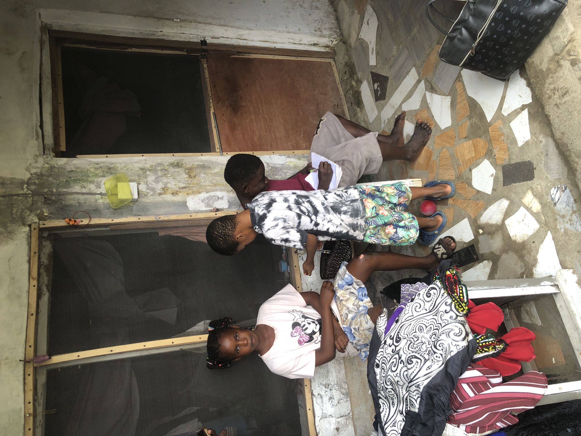 In Ado, Nigeria, a young girl with braided hair sits on a bench, wearing a pink t-shirt with a Minnie Mouse graphic and a floral skirt. To her right, a boy in a tie-dye t-shirt and patterned shorts stands, seemingly handing something to an older boy. The older boy, seated on the ground, wears khaki shorts and a red shirt and is writing on a notepad. In the foreground, a pile of clothes, including a black and white patterned garment and red and striped items, is stacked on a white metal structure. The scene unfolds on a tiled floor in front of a building with a window covered by mesh and a wooden door. A black bag rests on the floor to the right.