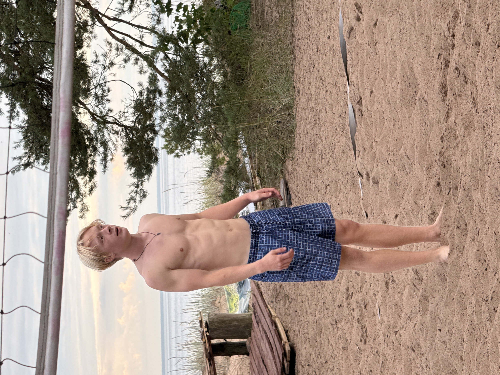 A young, shirtless Caucasian male with blonde hair stands on a sandy beach, facing the right. He is wearing blue checkered swim trunks and a dark necklace. In the background, a body of water can be seen, with a volleyball net strung across the top of the frame and a pine tree to the right. A wooden structure is visible behind the male to the left. The setting appears to be Zelenaya Roshcha, Russia.