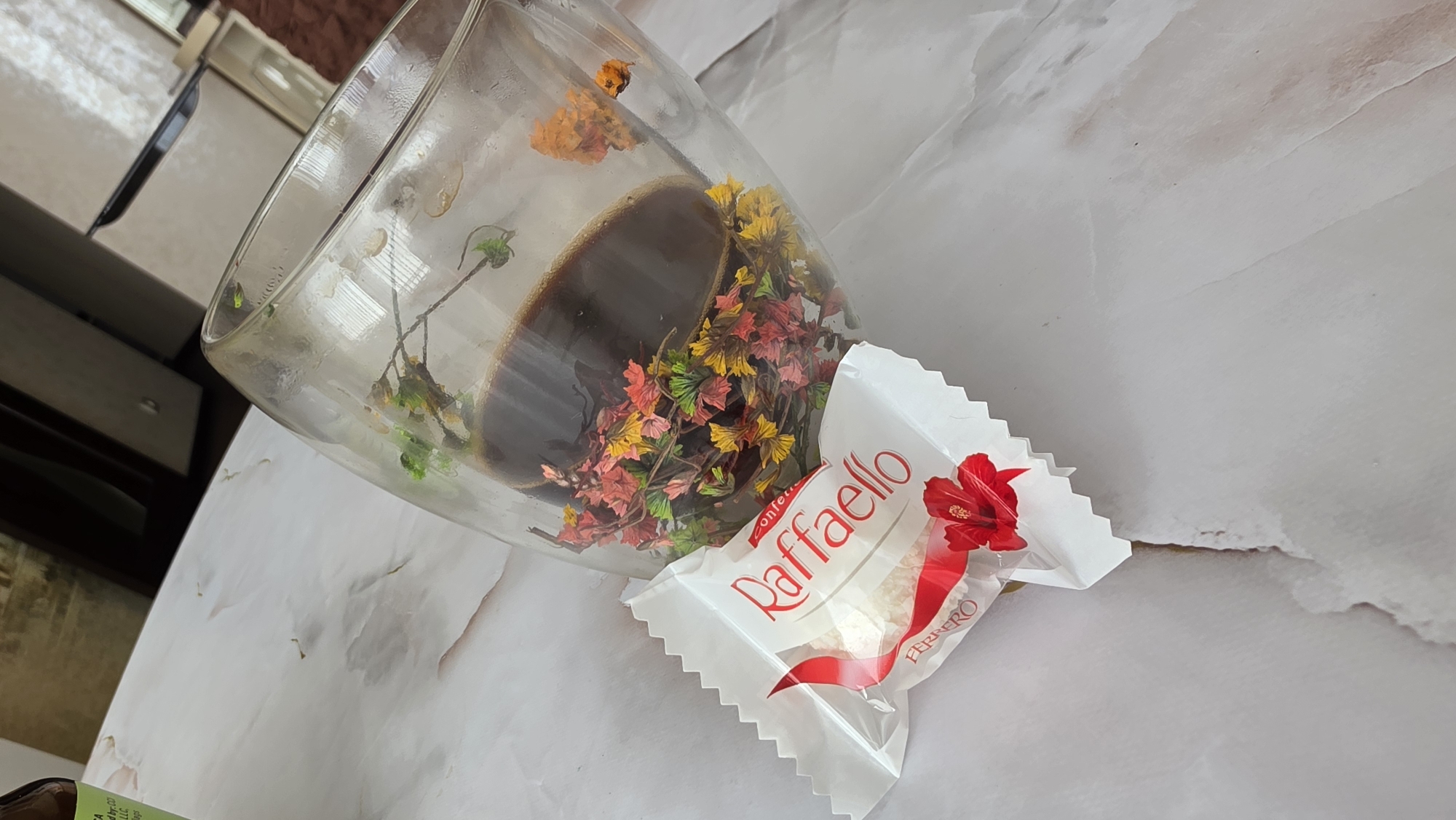 An overhead shot of a glass coffee mug filled with dark liquid and decorated with miniature artificial flowers is placed on a marble-patterned surface. To the left of the mug, a RaffaELLO candy in its wrapper lies on the surface. The candy wrapper is white with red lettering and a red hibiscus flower. The mug has a double-walled design and appears to be slightly tilted. The background is blurred, showing hints of indoor furniture.
