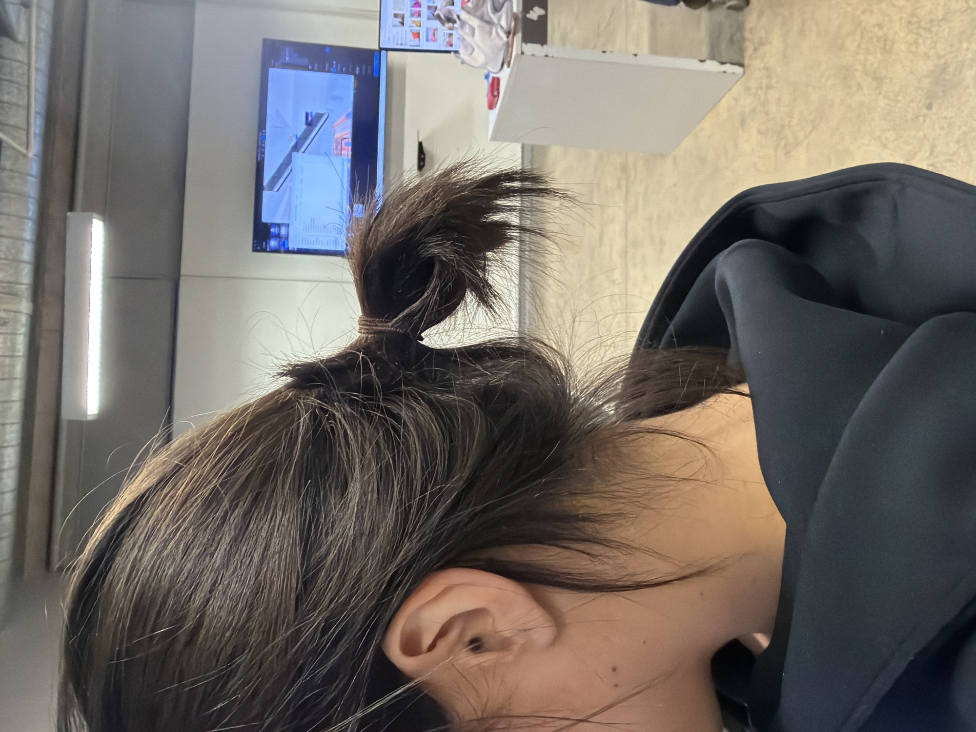 A person is depicted from the left side and rear, showcasing dark brown hair tied into a messy low ponytail with a dark elastic band. Loose hair strands are visible around the head and neck. The person's left ear is partially visible, containing a small, dark circular object. Two small moles are present on the person's lower neck. The subject is wearing a dark, possibly black or navy blue, hooded garment.
The scene is an indoor, well-lit environment with light-colored walls and an industrial-style ceiling featuring exposed ducts and a long, rectangular LED light fixture. In the background, a large flat-screen monitor displays 3D design software with architectural or structural elements in light blue and grey tones. Below and to the right of this, a second, smaller monitor shows a grid of multiple colorful thumbnail images. A white rectangular table or partition stands to the right, supporting a red object and a light-colored, crumpled object. The floor appears to be a light-colored, possibly concrete, surface.
The location is an interior space, likely an office, design studio, or educational facility, situated in Mexico City, Mexico.