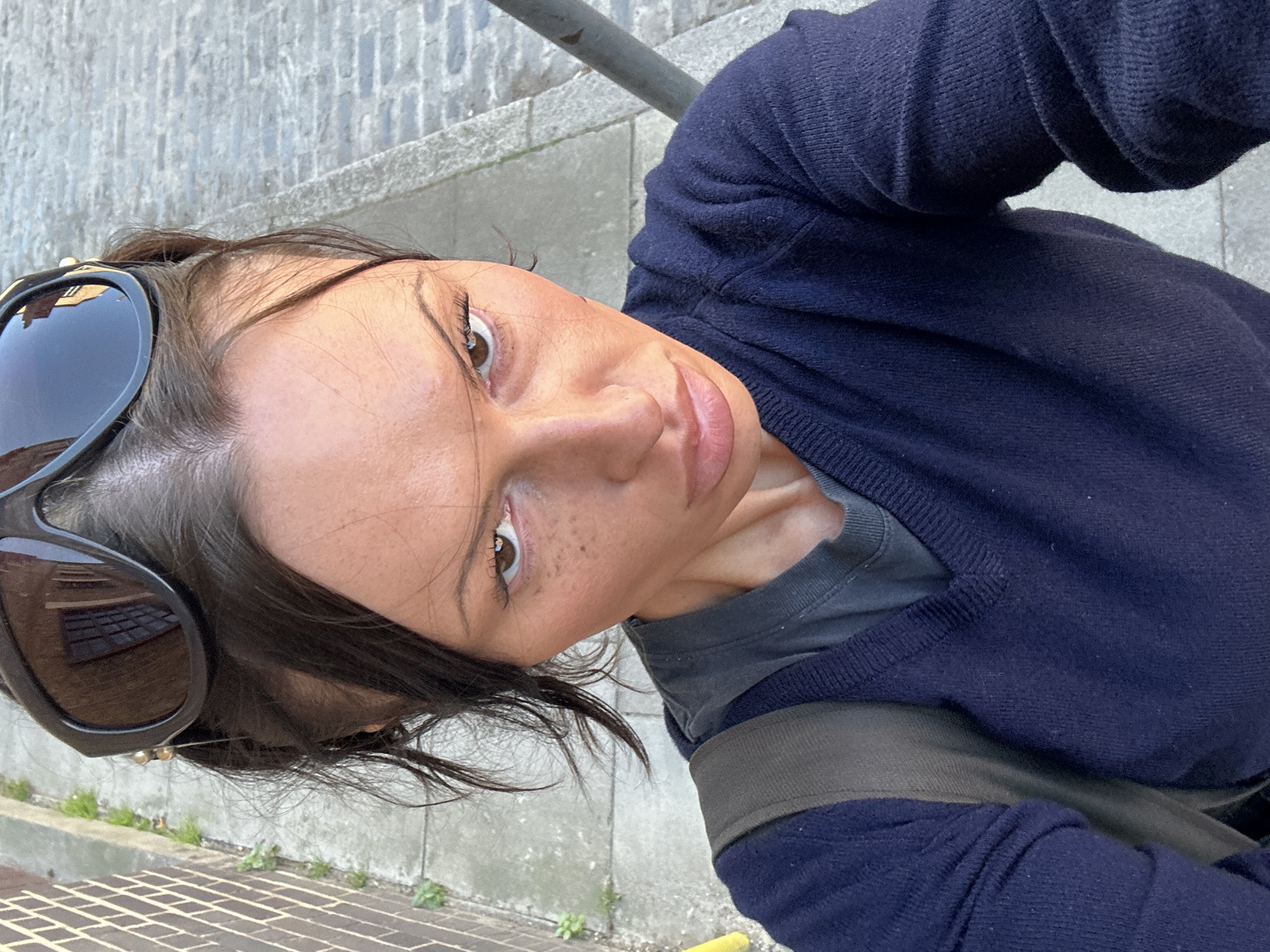 An eye-level outdoor portrait features a person with dark hair, a medium skin tone, and brown eyes, looking directly at the camera with a neutral expression. They have some freckles visible on their nose and cheeks. Dark-lensed sunglasses are pushed up onto their head.
The person is wearing a dark navy blue knitted cardigan or sweater over a grey t-shirt, with a dark olive-green or khaki strap from a bag visible across their shoulder. The image appears to be a selfie, taken from a slightly low angle, showing their upper body and face.
The background suggests an urban setting, possibly in London, United Kingdom, with grey stone paving (resembling cobblestones or square pavers) and a concrete curb. A metal railing is visible behind them. Small patches of green vegetation are growing along the curb. The lighting indicates it is daytime, with soft, diffused light. There is no visible text in the image.