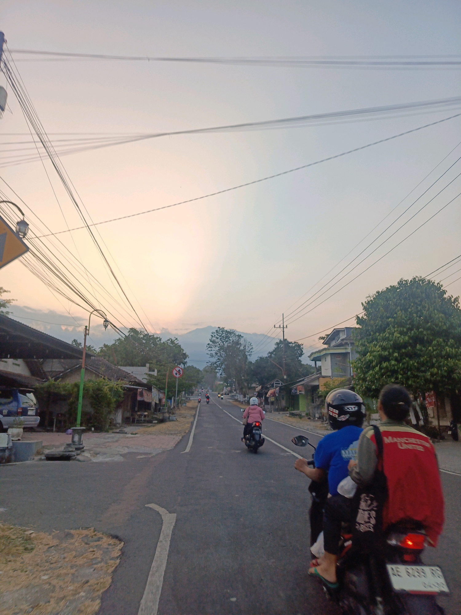 The image captures a scene on a paved road in Kalang, Indonesia, during what appears to be dusk. The sky is a pale orange and blue gradient, with visible power lines crisscrossing overhead, casting faint shadows. On the right side of the road, there are several houses with tiled roofs and some trees, one of which is a large, dense green tree. On the left side of the road, there are more houses, some with awnings, and a parked blue car. The road itself has a white line marking the lane.
Several motorcycles are present. In the foreground, two people are riding on a motorcycle, with the person in the back wearing a red jacket with "MANCHESTER UNITED" printed on it, and the person in front wearing a blue shirt and a black helmet. Another motorcycle with a rider wearing a pink jacket is ahead of them, traveling in the same direction. Further down the road, more motorcycles are visible, indicating traffic. A traffic sign with a red circle and a diagonal bar is also visible on the right side of the road, suggesting a restriction. The overall impression is a quiet, everyday scene in a rural or suburban area of Kalang.
