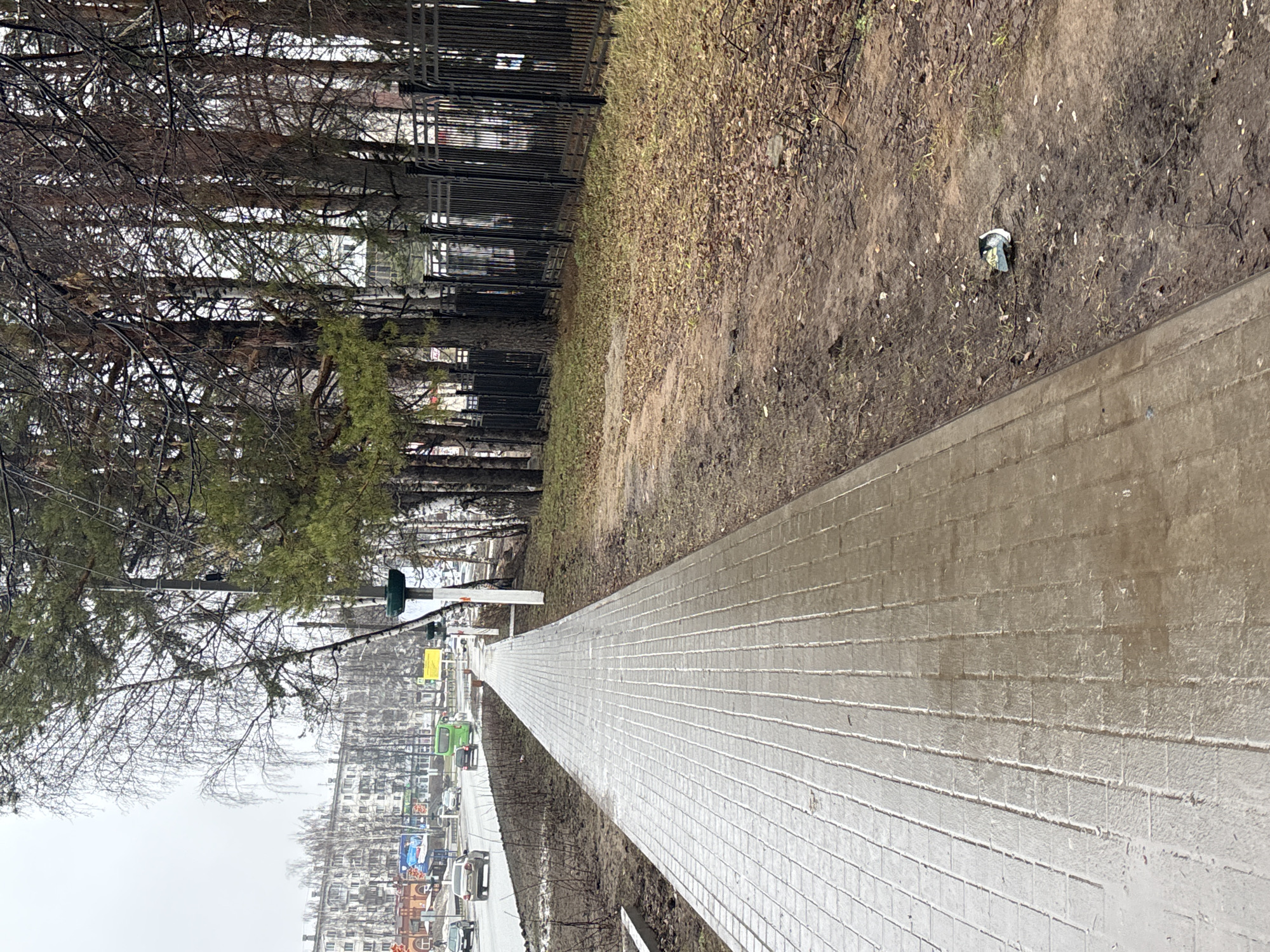The image depicts an outdoor scene in the city of Chaikovsky, Russia. A pedestrian walkway constructed of light gray, textured rectangular pavers stretches lengthwise into the distance, narrowing due to perspective. To the left of the walkway, a road is visible with several cars and a green municipal vehicle. Beyond the road, buildings are present. To the right of the walkway, there is a strip of dirt and dead leaves. A discarded piece of trash is on the dirt. A line of trees runs along the left side of the image. A black metal fence is positioned next to the trees. The weather appears overcast.