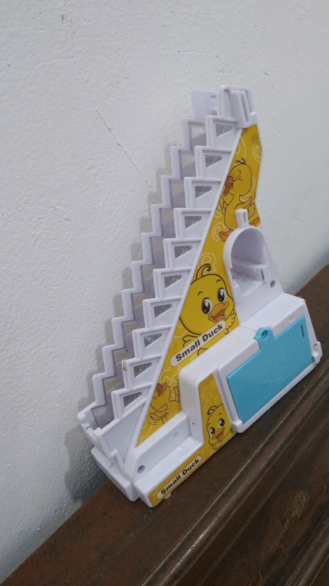 The image shows a toy duck-themed sliding staircase on a wooden surface against a white textured wall. The toy is white with yellow accents featuring cartoon ducks and the text "Small Duck." It has multiple tiered steps and a blue compartment on the side. The toy is positioned at an angle, with its base resting on the wooden surface. There are no people or actions depicted in the image. The environment appears to be indoors, possibly a room in Kebon IX, Indonesia, though this location cannot be definitively confirmed from the visual information alone.