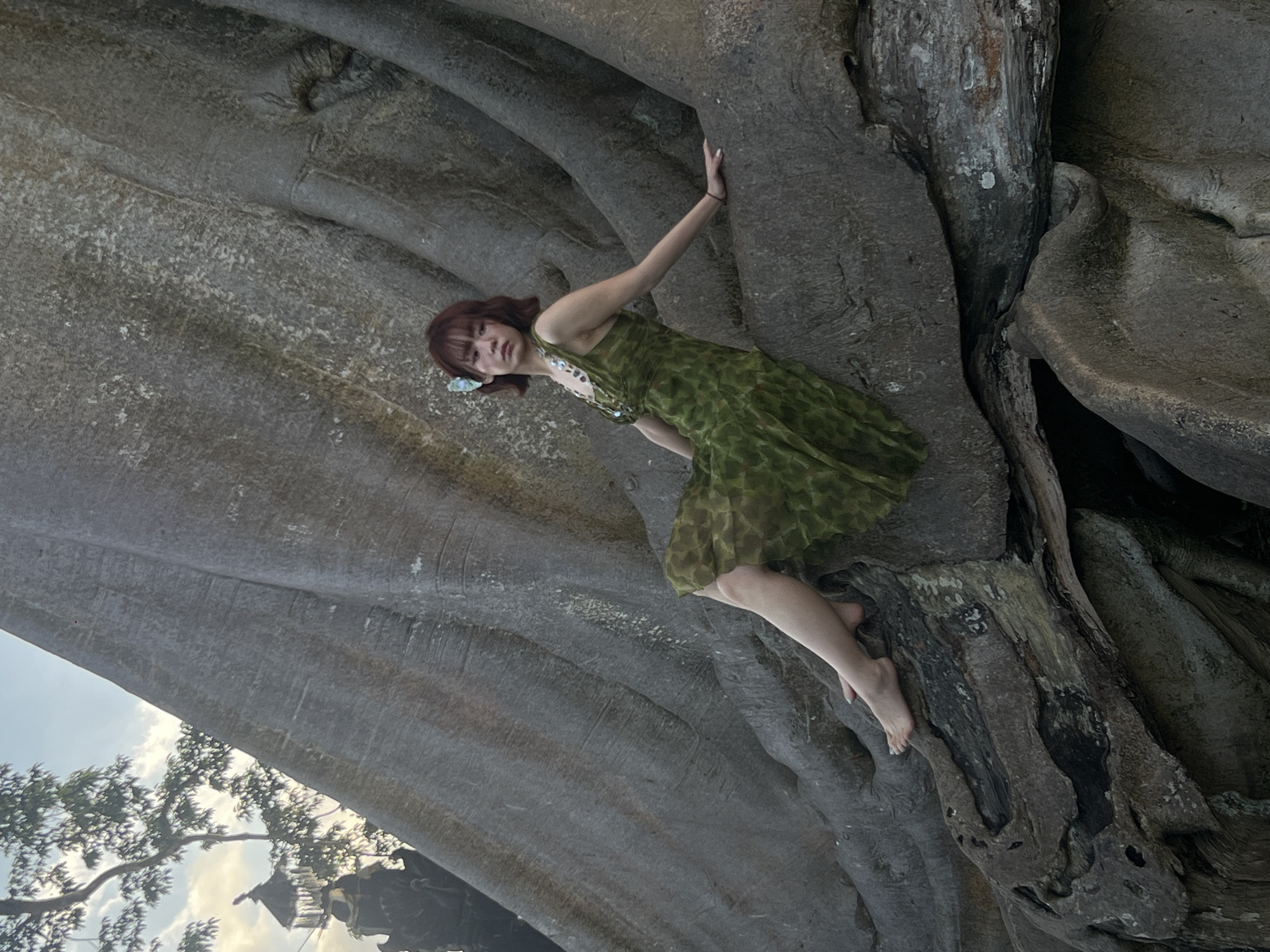 A young woman with short, dark hair adorned with a light blue flower sits on the exposed roots of a large tree. She is wearing a sleeveless, green dress with a subtle patterned texture. Her left leg is extended, and her right leg is bent. Her left arm rests on her knee, and her right arm is extended back, with her hand resting on a root. The tree's roots are massive and textured, appearing weathered and ancient. In the background, partially obscured by the tree and foliage, is a glimpse of traditional Balinese architecture, specifically a tiered umbrella structure, suggesting the location is in or around Marga, Indonesia. The sky is visible in the upper left portion of the image, with soft clouds and the muted light of either dawn or dusk. The overall scene has a serene and slightly mystical atmosphere.