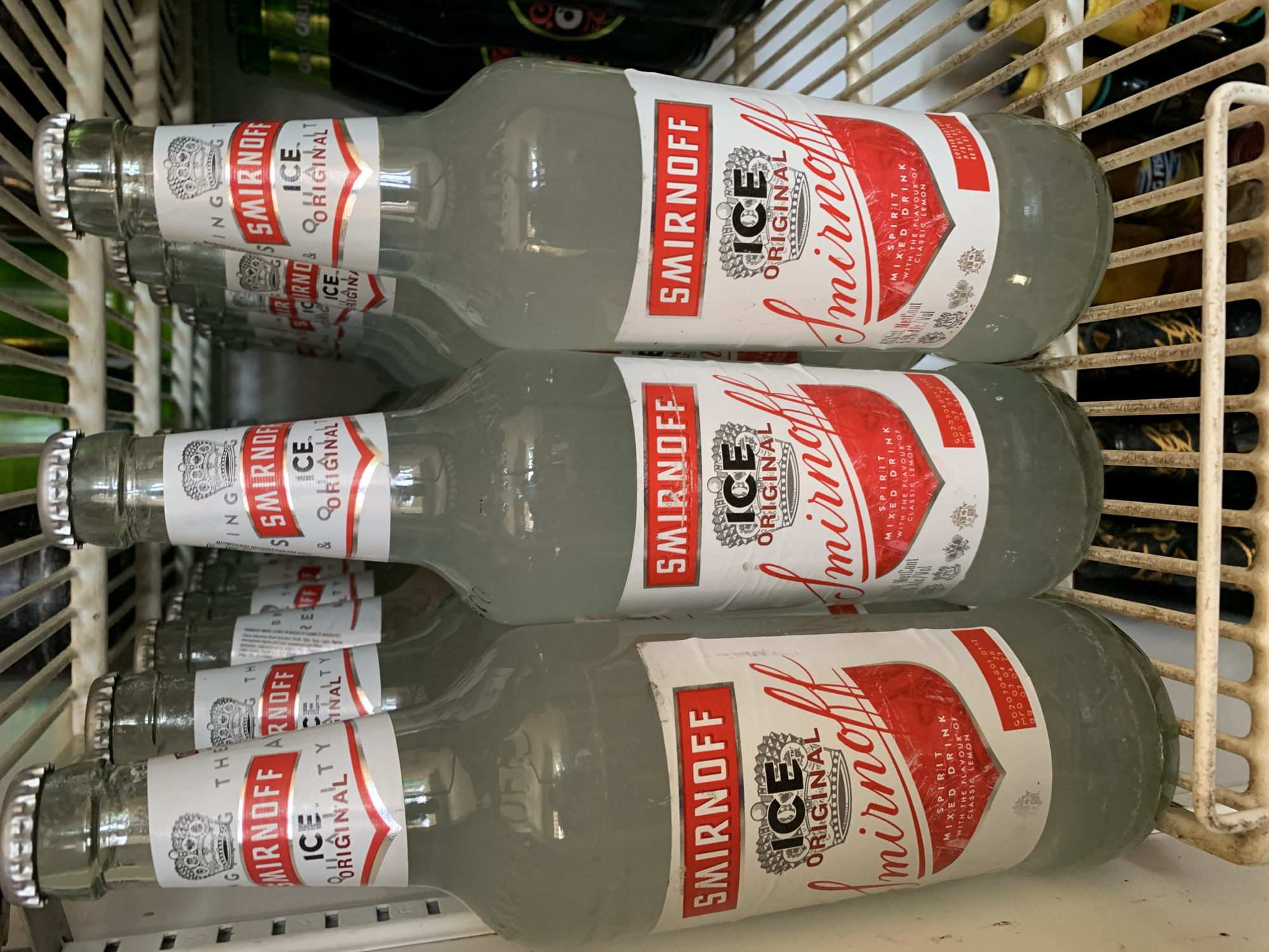 The image shows a wire basket containing six bottles of Smirnoff Ice Original. The bottles are horizontally stacked, with their labels facing upwards and to the right. The bottles are transparent and appear to contain a clear liquid. The labels are predominantly white with red and black lettering. The basket's wire frame is visible and partially obscures some of the bottles. The location and any human subjects or actions are not discernible from the image.