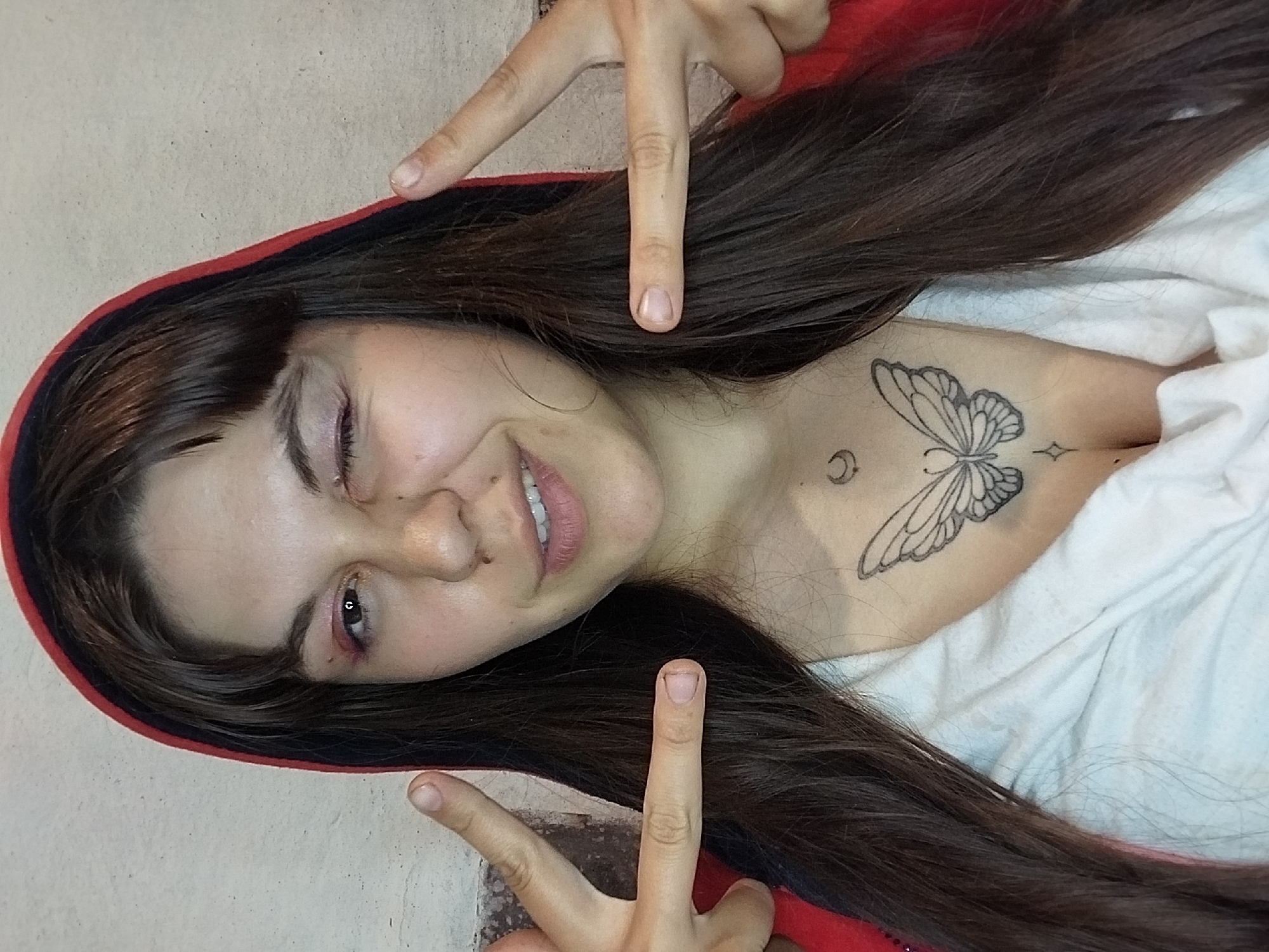 A woman with long, dark hair and a red hooded jacket is looking at the camera, winking and holding up peace signs with both hands. She has a butterfly tattoo on her chest. The background appears to be a textured wall.