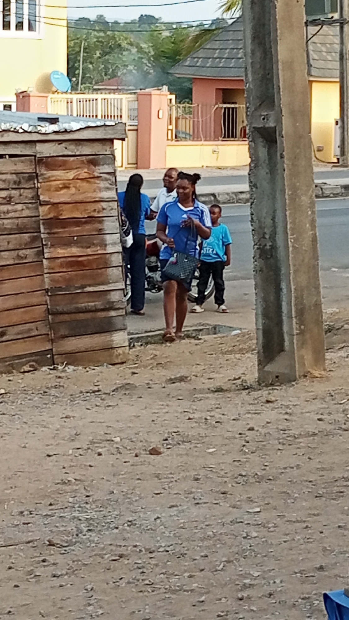 A group of people are on a street in Adankolo, Nigeria. In the foreground, there is a dirt and gravel surface. To the left, there is a structure made of wooden planks. A woman in a blue shirt and dark skirt is walking forward carrying a handbag. Behind her, there is a woman with long braids wearing blue and jeans, a man in a white shirt, and a child in a blue shirt and black pants. A parked motorcycle is near them. In the background, there are buildings, some with tiled roofs, and some trees. An electrical pole is to the right.