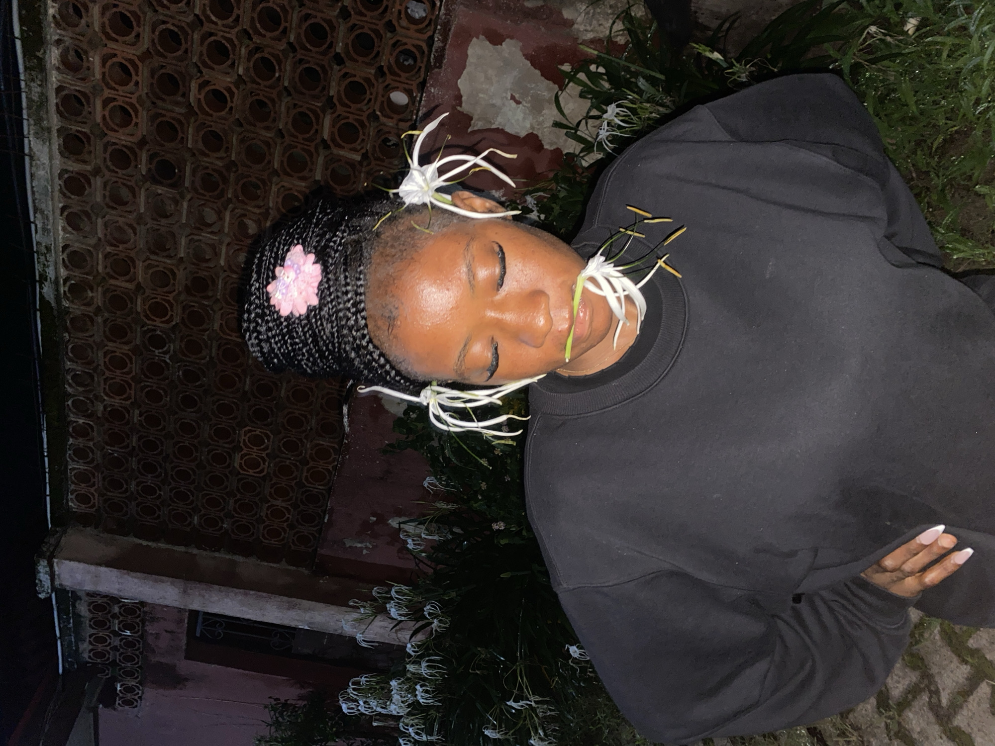 A medium shot captures a young Black woman with her eyes closed, adorned with white spider lilies on her braided hair and in her mouth, standing outdoors at night. She wears a black crew neck sweatshirt and has her left hand resting on her hip. Behind her, a textured, dark red brick wall with circular openings runs along the top of the frame, partially obscured by a reddish-pink building on the left. Lush green foliage, also dotted with white spider lilies, fills the mid-ground and foreground. The scene appears to be in a residential area of Jos, Nigeria, with the characteristic architectural style of the wall and building suggesting a local setting.