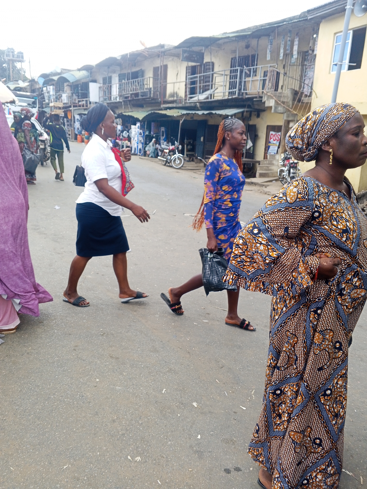 This is a medium-shot, eye-level, outdoor photograph taken on a street in Okene, Nigeria, likely in a market or commercial area. The foreground is dominated by the lower half of a woman wearing a colorful, patterned maxi dress and a matching headscarf. She has on sandals and is holding a black bag in her right hand. To her left, a woman in a blue, patterned dress with her hair in braids is walking away from the camera, also carrying a black bag. Further to the left, a woman in a purple garment is partially visible. In the background, several other people are walking and interacting, and there are buildings lining the street, suggesting a busy urban environment. Motorcycles and a white vehicle are also present. The street is paved and appears somewhat worn. The sky is overcast.