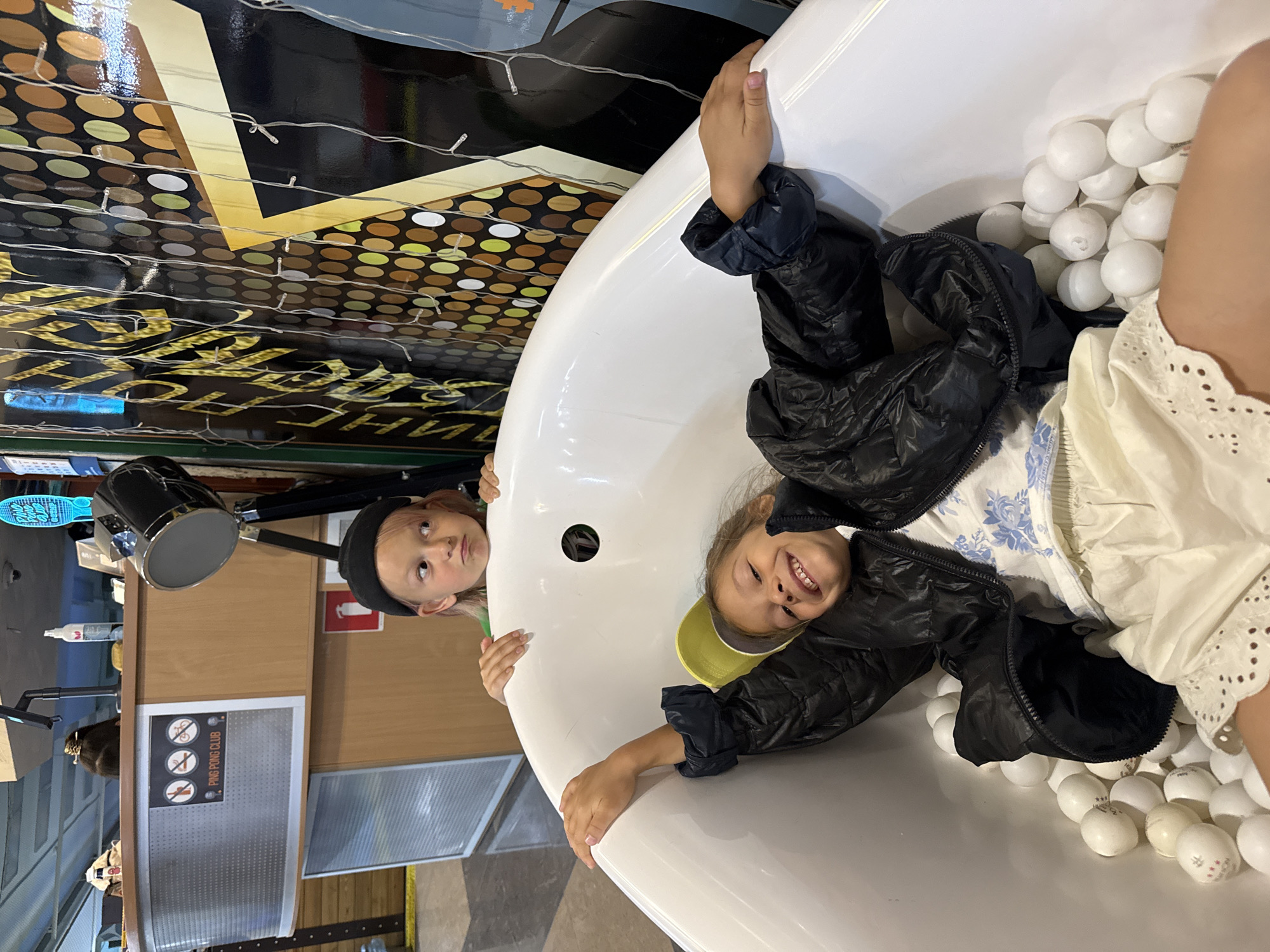 The image depicts two young children inside a white bathtub filled with white ping pong balls. The child in the foreground is lying down, smiling and holding onto the edge of the tub. They are wearing a black puffy jacket, a white and blue floral shirt, and light-colored shorts with eyelet trim. A yellow baseball cap is visible. The second child is behind the first, leaning over the edge of the tub and looking to the side with a slightly annoyed expression. This child is wearing a black beanie. The bathtub appears to be situated in an indoor space with a counter and a wall decorated with colorful circles and string lights in the background. A sign with "PING PONG CLUB" and crossed-out icons is visible on the counter. The overall impression is a playful and informal setting.