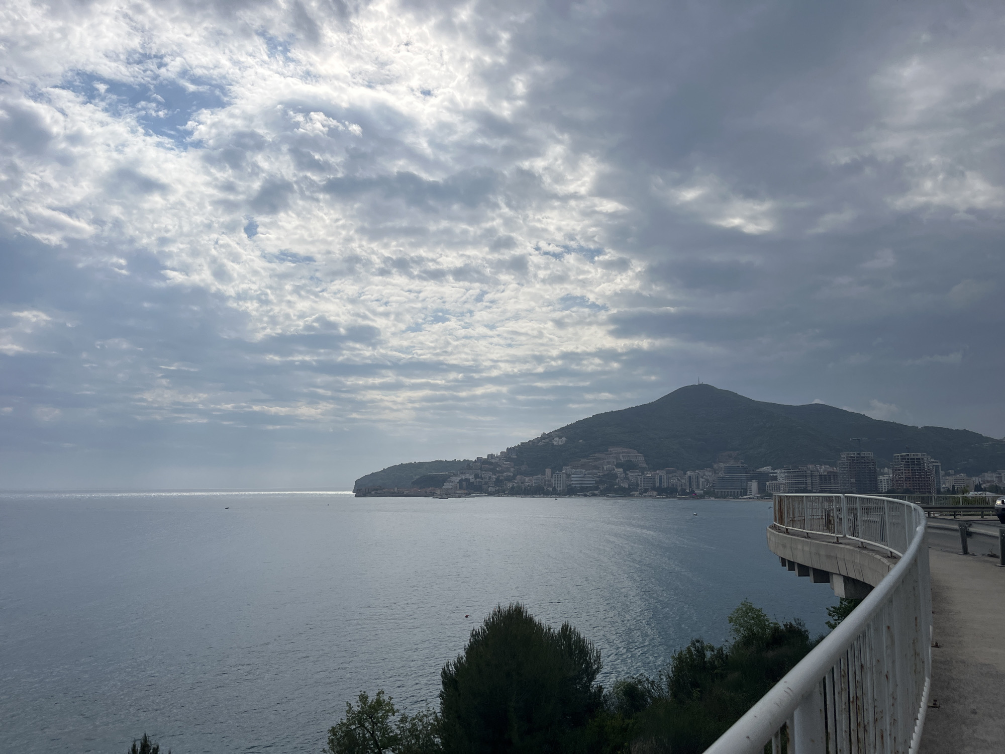 This panoramic photograph captures a scenic coastal landscape in Budva, Montenegro, under a dynamic sky. In the foreground on the right, a curved white metal railing defines an elevated pathway or road, with green foliage and trees visible below.
The vast expanse of the sea dominates the middle ground, its surface reflecting glints of sunlight that occasionally break through the cloud cover. The water appears calm, extending towards a hazy horizon.
In the background, a prominent mountain rises, its lower slopes and base densely populated by the coastal town of Budva. Numerous buildings, including hotels and residential structures, are visible along the shoreline and ascending the mountain, showcasing urban development integrated with the natural terrain.
The sky is largely overcast with a mix of grey and white clouds, suggesting a cloudy but bright day. Patches of blue sky are discernible between the clouds, especially towards the upper left. The overall lighting is diffused, contributing to a serene yet expansive atmosphere. No people or visible text are present in the image. The scene depicts a vibrant coastal city nestled against a mountainous backdrop, viewed from a scenic overlook.
