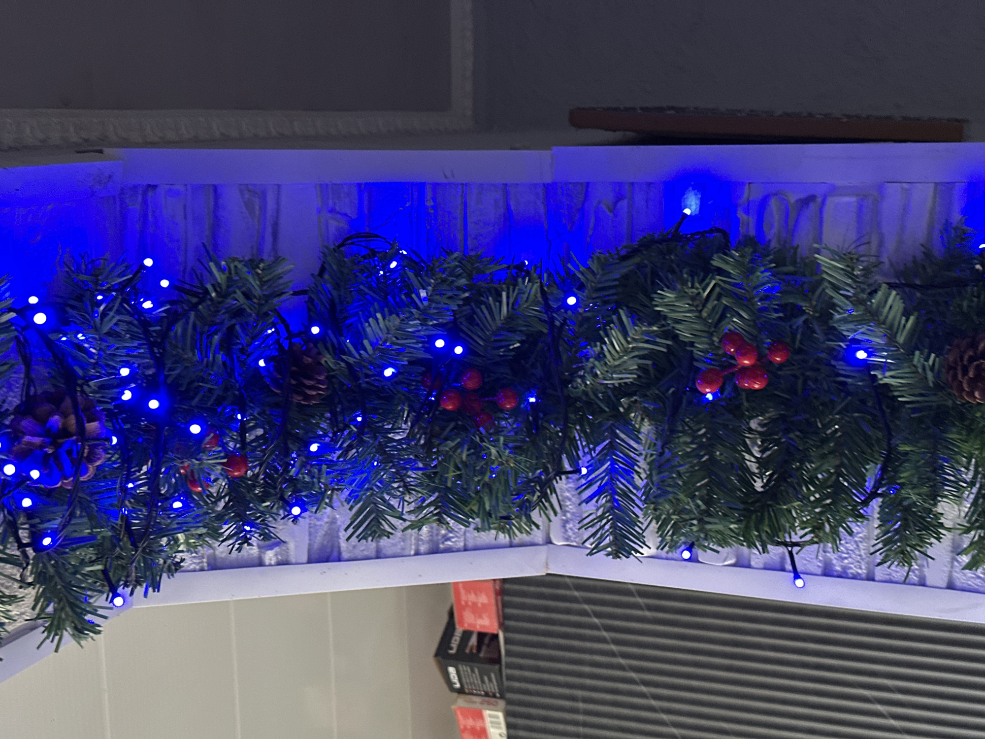 A forensic image depicts a vertical segment of artificial evergreen garland, illuminated by numerous blue LED string lights. The garland is further embellished with several clusters of red artificial berries and scattered brown pinecones. This decorative element is affixed to a white, vertical architectural trim or frame, which is positioned against a textured wall, possibly white brick or stone, receiving a strong blue cast from the lights.
To the left of the garland, the upper portion of the frame transitions to a light-colored, horizontal ceiling or paneling. Below this, a dark, corrugated or ribbed surface is visible, above which multiple cardboard boxes are stacked. Some boxes display partial text such as "LIDE" and "UDB". To the right of the garland, beyond the white trim and textured wall, a darker, unadorned wall section is present, bordered by a thin, vertical brown wooden element. The overall scene is dimly lit, with the blue LED lights serving as the primary illumination source. The location is unknown, Unknown.