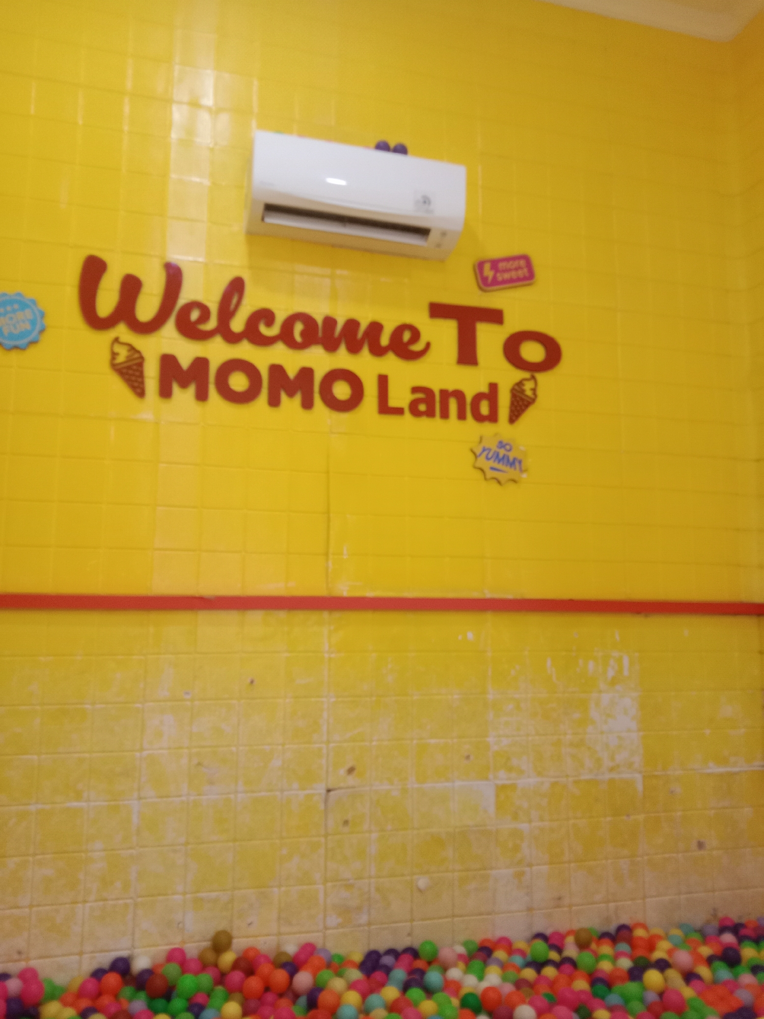 The image depicts a bright yellow tiled wall with "Welcome To MOMO Land" written in red, stylized lettering. An air conditioner unit is mounted high on the wall. Below the text, there is an image of an ice cream cone and a sticker that says "SO YUMMY". To the left of the main text, there's a blue circular sticker that appears to say "MORE FUN". The bottom of the image shows a ball pit filled with colorful plastic balls. This scene is located in Madiun, Indonesia.