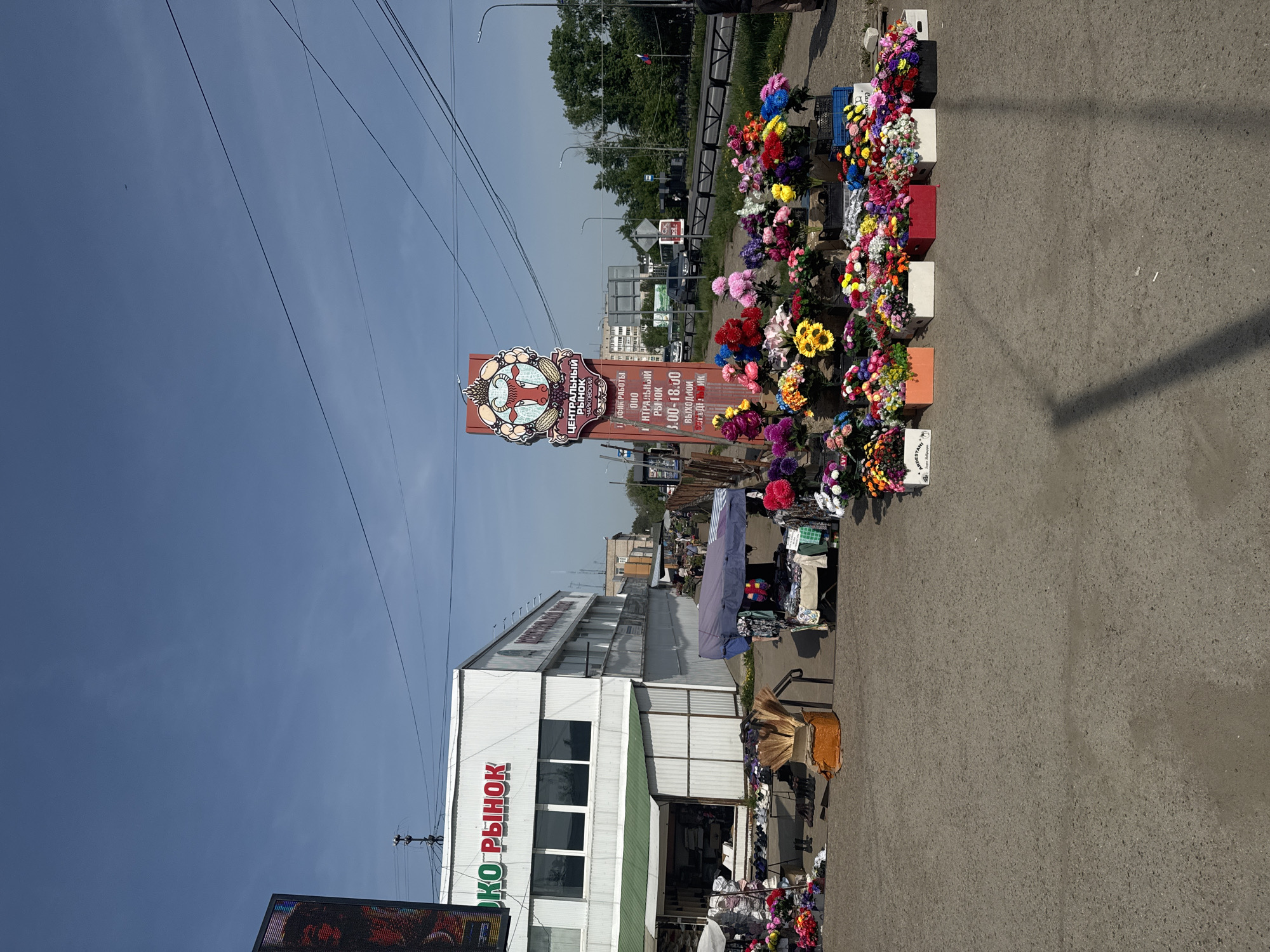 The image depicts an outdoor market in the city of Chaikovsky, Russia. A sign for "Centralny Ryznok" (Central Market) is visible, and another, larger sign identifies it as the Central Market of Chaikovsky, providing the hours of operation. Rows of artificial flowers in containers line the edge of the roadway. The market building is white with green accents. People are present but indistinct within the market area. Overhead power lines are visible against a blue sky.
