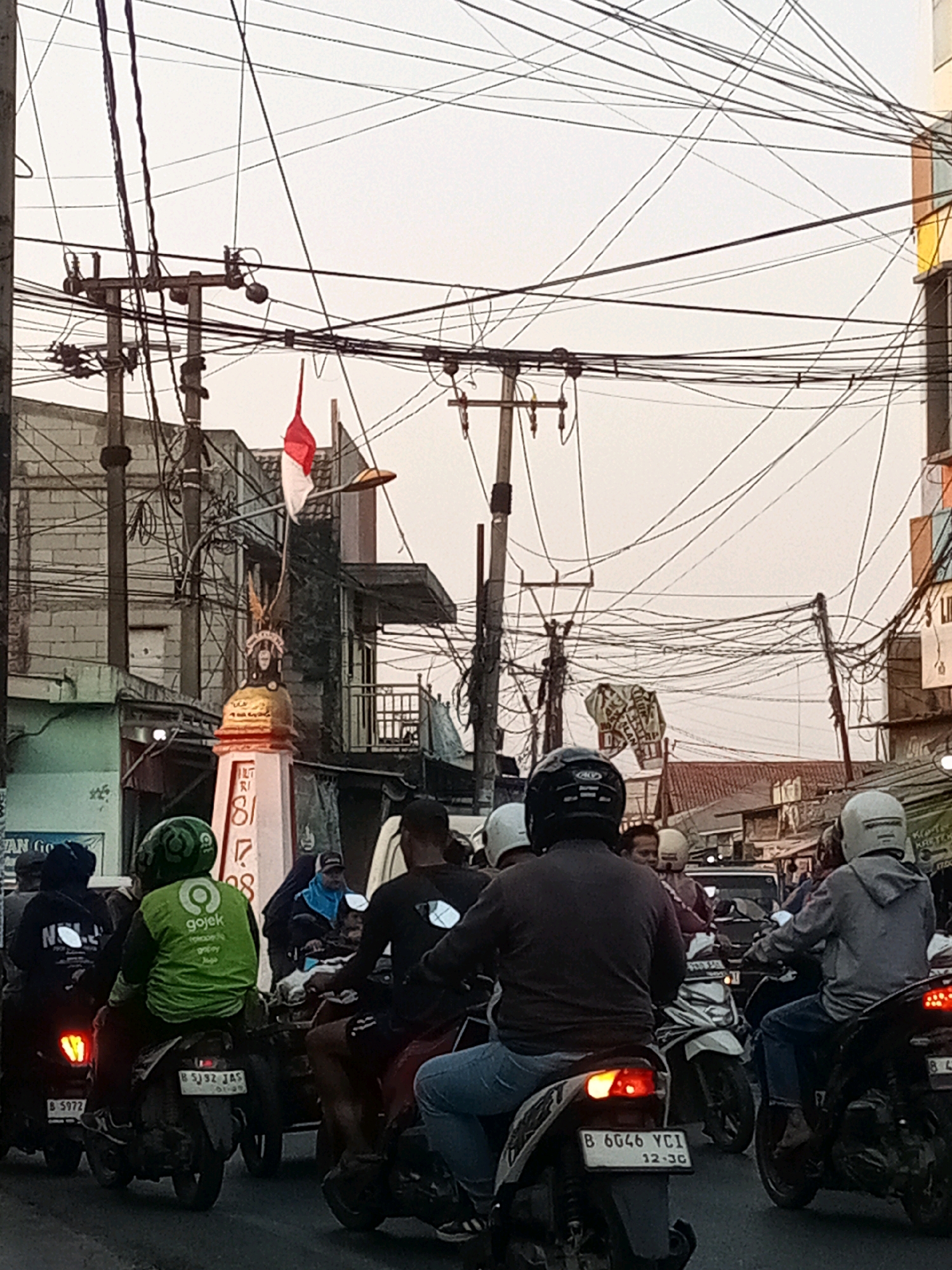 In Karangsatria, Indonesia, a street scene depicts heavy traffic on a cloudy day, with numerous motorcycles and their riders navigating the road. Prominently featured is a monument adorned with a red and white flag, likely the Indonesian national flag, positioned near a power pole tangled with a dense network of electrical wires. Several individuals are seen on motorcycles, many wearing helmets for safety. One rider is wearing a bright green Gojek jacket, indicating a ride-hailing service. Buildings line the street, some appearing to be shops or residences, and a variety of signs are visible, though not all are legible. The overall atmosphere suggests a bustling urban environment with organized chaos characteristic of busy Indonesian city streets.
