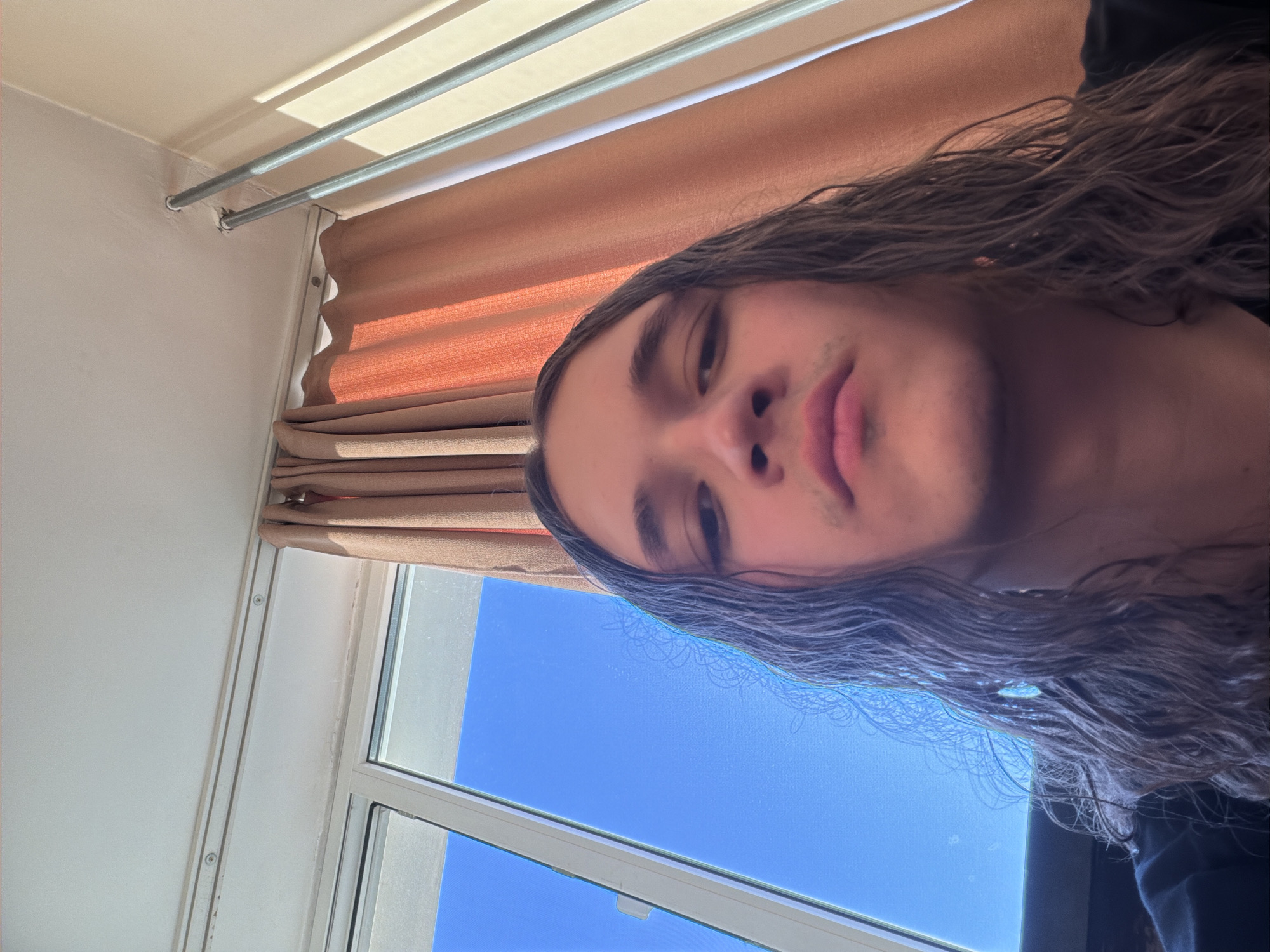 The image captures a close-up, low-angle shot of a young person with long, wavy dark hair and light facial stubble on their upper lip and chin. Their head is tilted, presenting their face horizontally across the frame with a neutral expression as they look towards the camera.
The setting is an indoor room, likely in Plovdiv, Bulgaria, brightly lit by natural light. A white-framed window is visible in the lower left, revealing a clear, vibrant blue sky, indicating daytime and good weather. Partially drawn, light-colored pleated blinds or curtains hang over the window. High on the wall near the ceiling, two parallel metallic rods are mounted, casting shadows on the white ceiling from the sunlight. The walls are also light-colored. No specific activity is taking place other than the person posing for the camera, possibly taking a selfie. There is no visible text in the image.