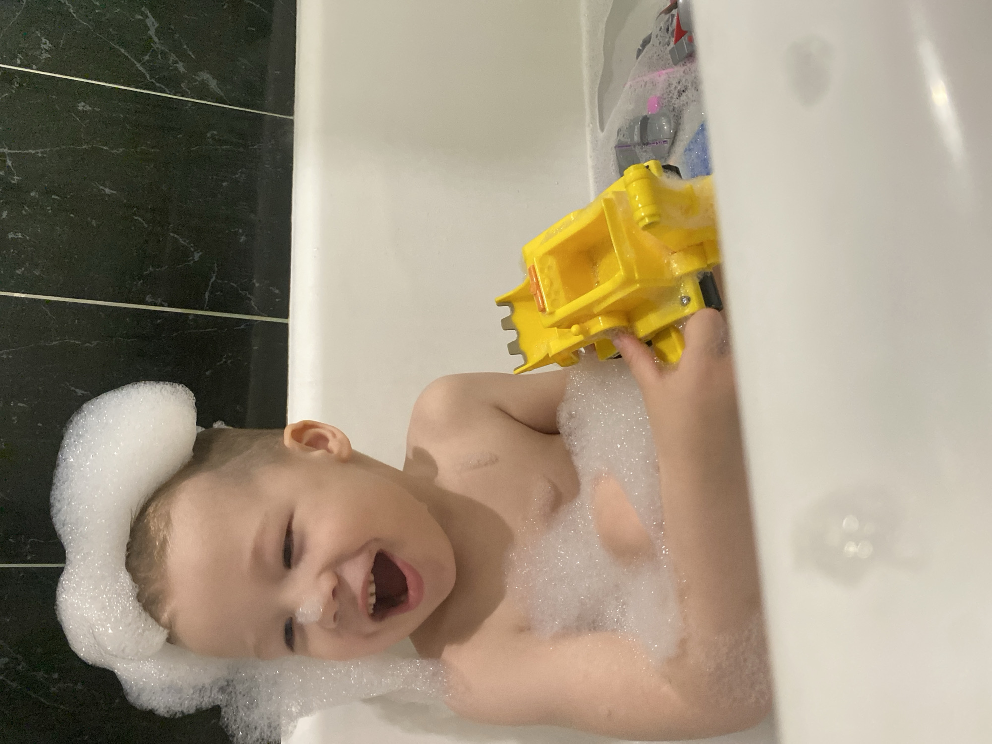 A young child is lying in a bathtub filled with bubbles. The child has bubbles on their head and body and is smiling and playing with a yellow toy truck. The bathroom has black tile on the wall. The city of Sochi, Russia, is not discernable in the image.