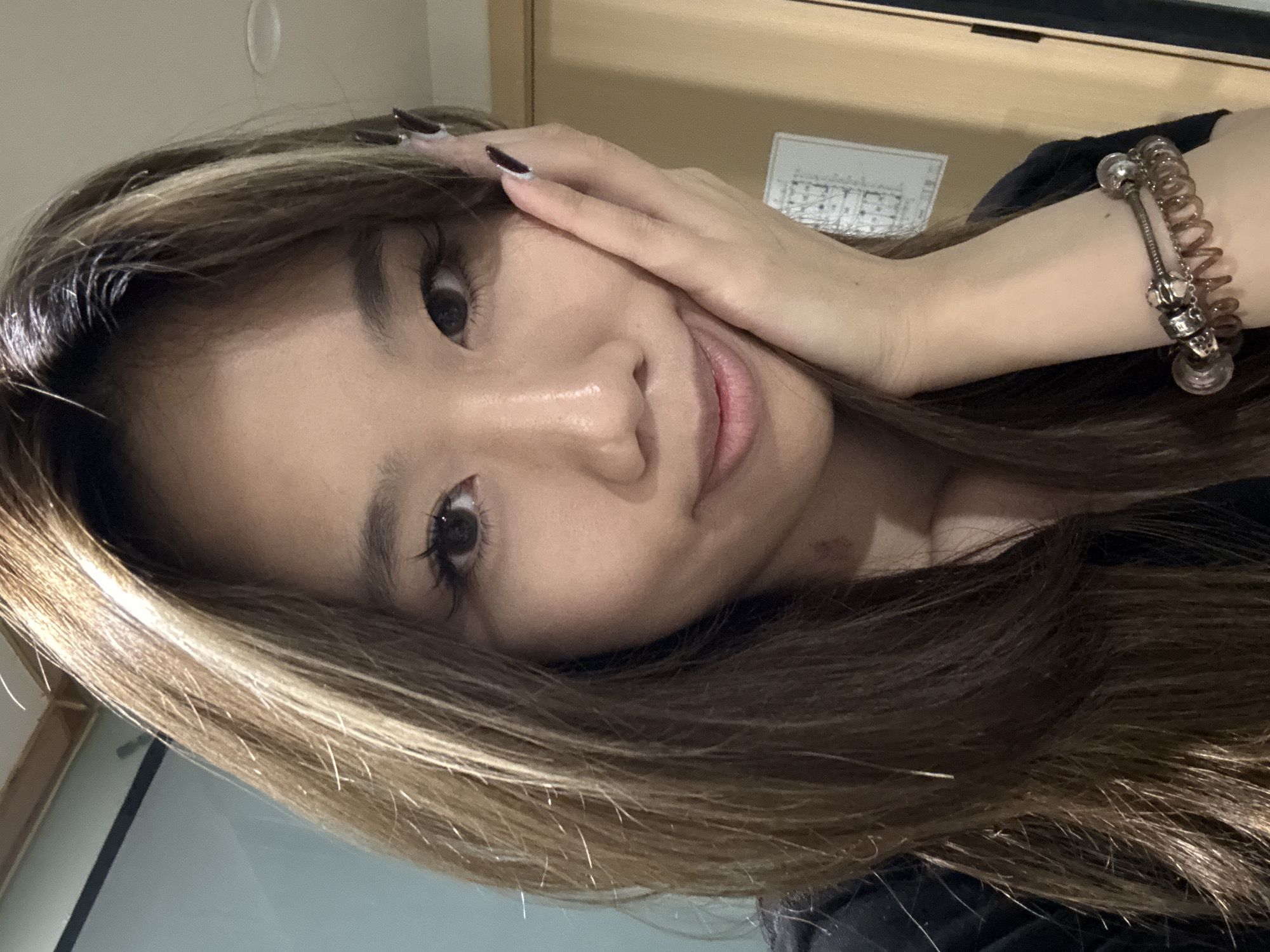 An Asian woman with light brown hair with blonde highlights is posing for a selfie. She is holding her hand to her face, with painted nails. She is wearing a bracelet. Behind her, there is a door and a calendar. There is no evidence of her location being Yuzhong District, China.