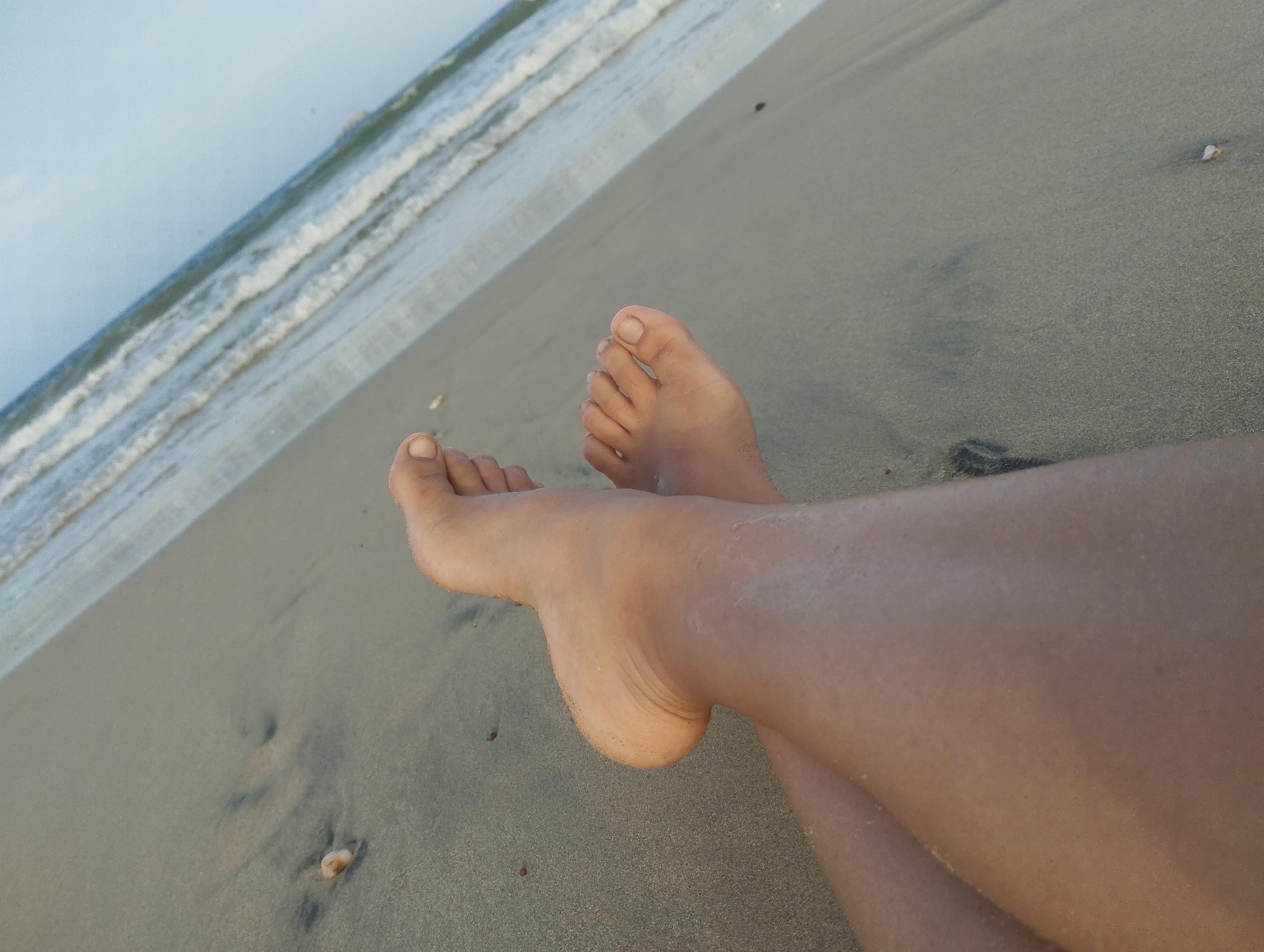 A close-up shot depicts a pair of human legs resting on a sandy beach. The feet are positioned towards the top center of the frame. Behind the legs, waves from the ocean can be seen meeting the shore. The scene appears to be located at a beach in Porlamar, Venezuela, given the context provided. The sand has a dark, wet appearance.