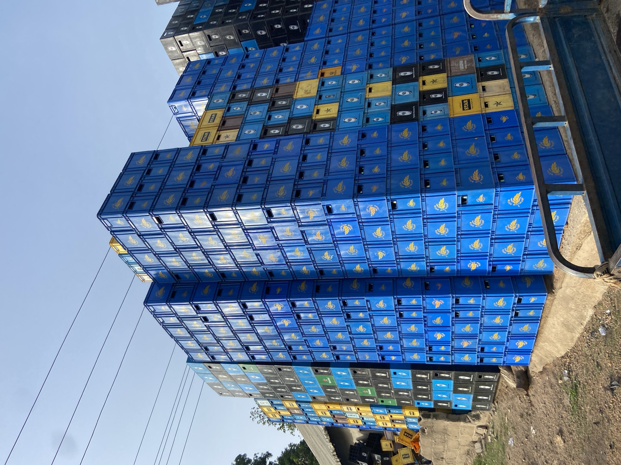 This media file presents a massive outdoor storage area filled with towering stacks of plastic beverage crates, predominantly in blue, black, and yellow colors, situated in Mararaba, Nigeria. The crates are piled many layers high, forming imposing, wall-like structures that reach upwards towards a clear, bright blue sky.
The majority of the visible crates are blue, each featuring a distinctive yellow logo: a stylized eagle or phoenix with spread wings, often associated with beverage brands. Closer inspection reveals the text "GULDER" printed on many of these blue crates. Interspersed within these blue stacks, and forming separate piles, are numerous black and yellow crates. Text such as "STAR," "LEGEND," and "GUINNESS" is clearly visible on these varied colored crates, indicating their use for different beverage products. Some crates also bear circular or oval labels.
The overall scene is static, with no human activity visible. Wires or cables are faintly discernible stretching across the upper portion of the frame against the sky. On the far right, a sturdy blue metal structure, possibly a railing, part of a loading ramp, or an industrial fixture, frames the edge of the crate stacks. The ground beneath the crates appears to be a mix of dirt and concrete, suggesting an informal storage yard or a commercial depot during daytime under clear weather conditions.