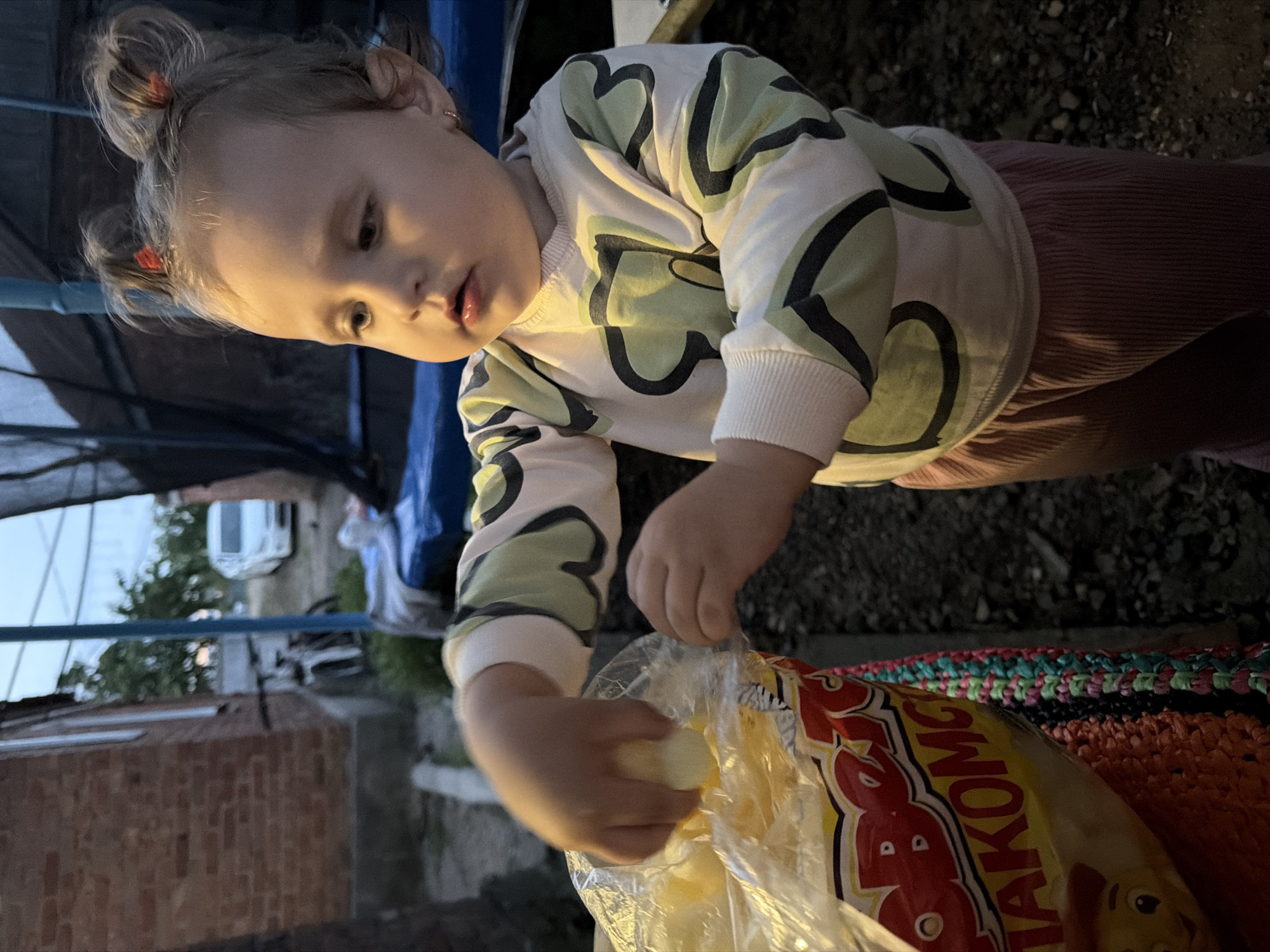 A young child, likely a toddler, is reaching into a clear plastic bag. The bag appears to contain some kind of food product, possibly snacks, with text visible on its red and yellow packaging. The child is wearing a light-colored long-sleeved shirt with a green and black pattern and pink ribbed pants. Their hair is styled in two small buns. The setting appears to be outdoors, possibly a backyard or an area near a building. In the background, a brick building, a bicycle, a white car, and a trampoline structure are visible, suggesting a residential area in Komsomolskiy, Russia. The lighting suggests it may be dusk or early evening.