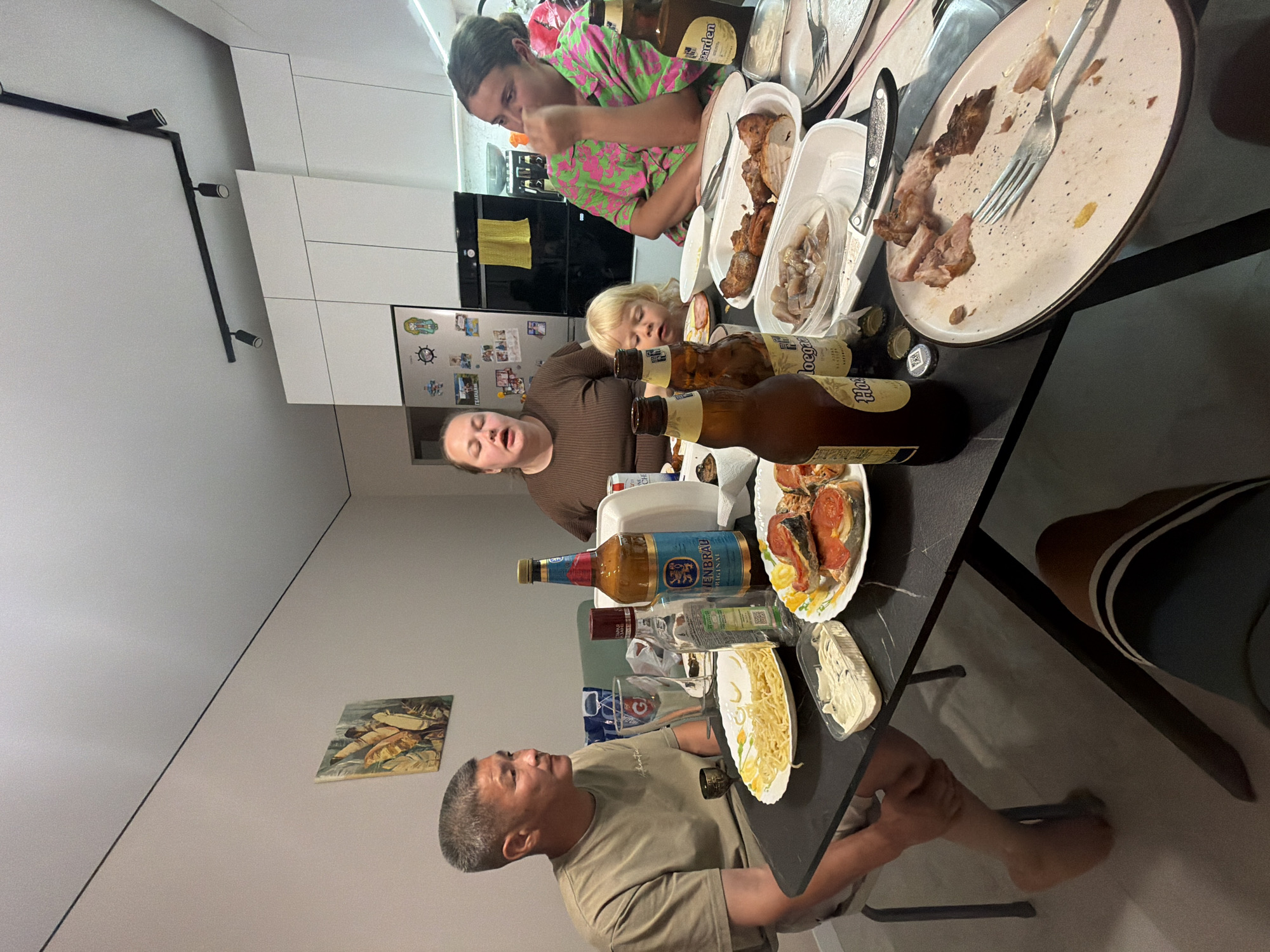Four people are gathered around a table in a kitchen or dining area. The table is laden with food and drinks, including several bottles of beer, a bottle of clear liquid, pasta, sandwiches with fish, and a container of dip. Two women, one with her mouth open as if speaking, and a young child are seated. A man is seated to the left of the table, looking to the right. A fourth person, mostly obscured, is seated on the right. The environment appears to be domestic, with a refrigerator and cabinetry visible in the background. The lighting suggests it is evening or indoors.
