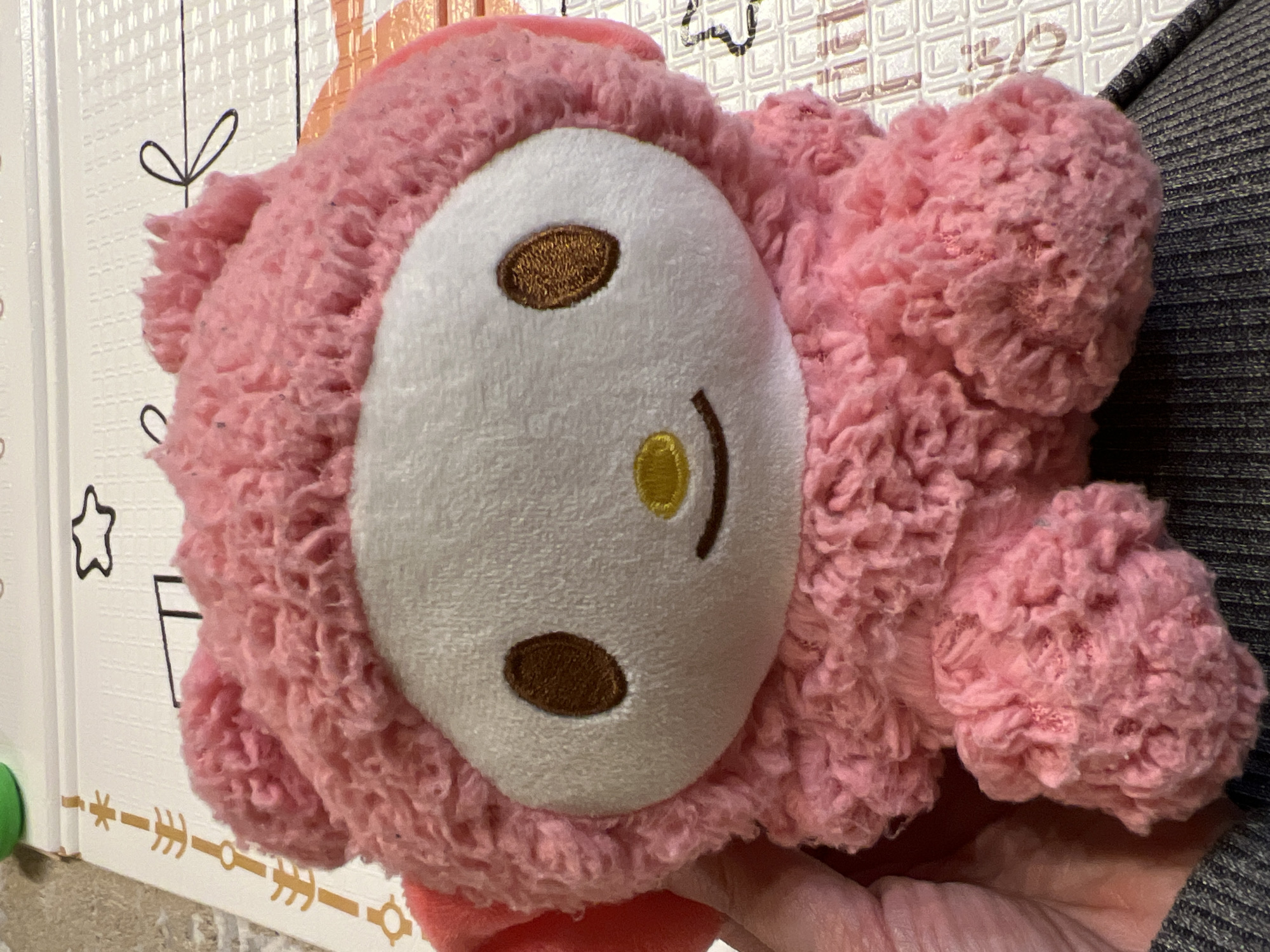 The image shows a pink, plush toy with a white oval face. The face has two dark brown oval eyes, a yellow dot nose, and a dark brown curved mouth. The toy has a fuzzy, textured body. The toy is being held in a hand. The background includes a patterned playmat with a black star and other black line designs on a white surface. There is also a dark gray fabric in the background on the right side.
The location in Yekaterinburg, Russia, cannot be determined from the given image.