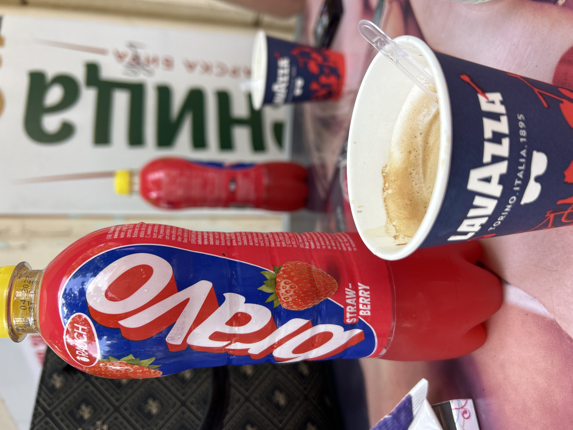 A high-angle shot captures a table setting featuring a bottle of Rauch Bravo Strawberry drink, a cup of Lavazza coffee, and some cigarettes. The bottle is red and blue with "Bravo" prominently displayed in large white letters. A strawberry graphic is visible on the bottle. The coffee cup, also blue and red, has "Lavazza TORINO.ITALIA. 1895" printed on it. The background shows a sign with Cyrillic lettering and another blurred bottle of Rauch Bravo. The scene is set outdoors on what appears to be a table with a pinkish, patterned surface. The lighting suggests daylight. The presence of a Rauch Bravo bottle with a Bulgarian inscription and the Cyrillic sign in the background strongly suggest the location is Obnova, Bulgaria.