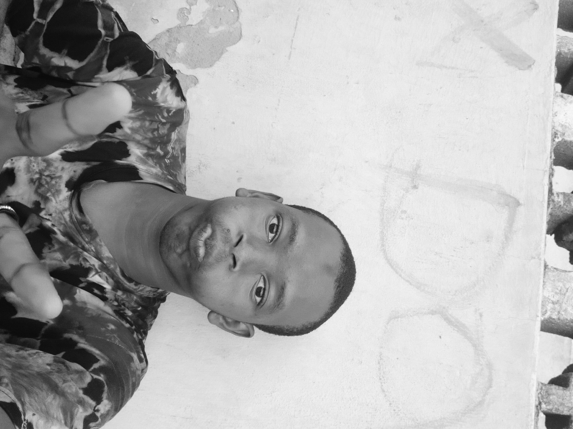 A black and white medium shot depicts a man with short-cropped hair in a patterned shirt standing against a textured, off-white wall. The wall has faded graffiti and patches of missing paint. The man is making a gesture with his left hand, holding two fingers out. The wall also appears to have a concrete structure to the right. Without additional information, the specific location in Lagos, Nigeria, cannot be determined.